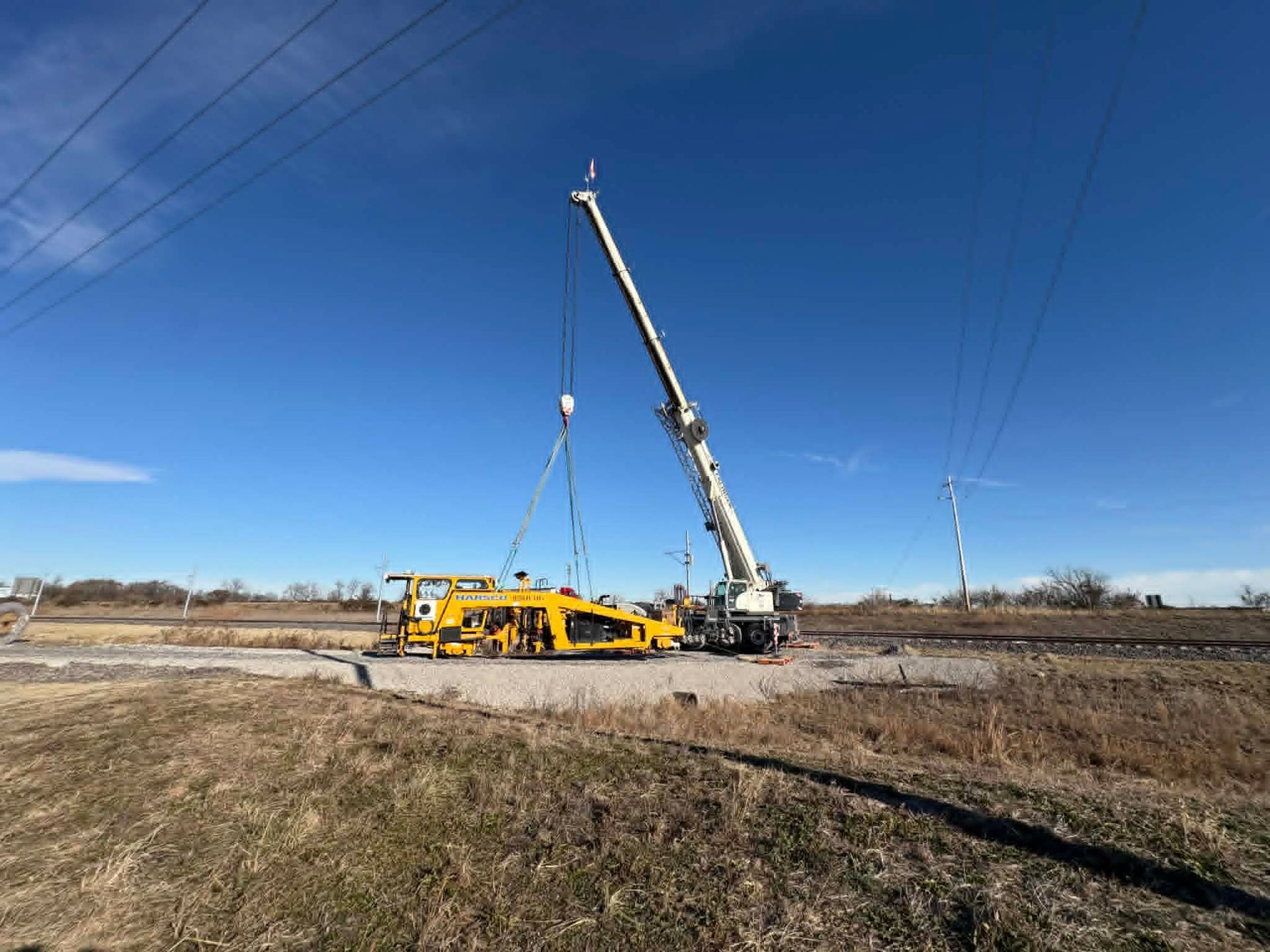 A crane lifting a yellow machine near railroad tracks under a blue sky.