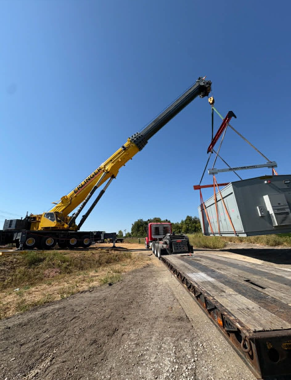 A large yellow crane lifting a gray industrial container off a flatbed trailer on a construction site.