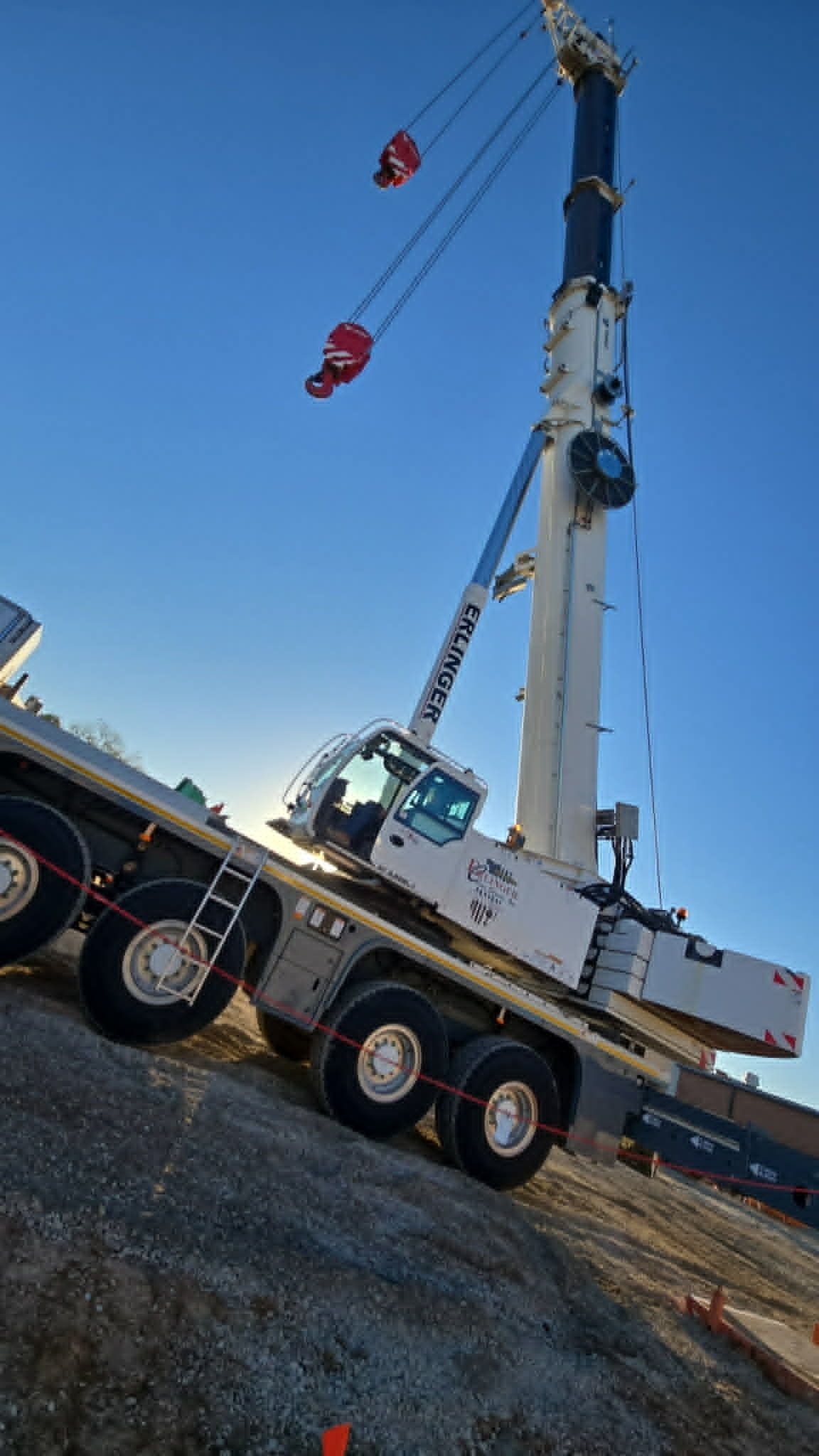 Large white crane on a gravel slope under a clear blue sky. Two red hooks hang.