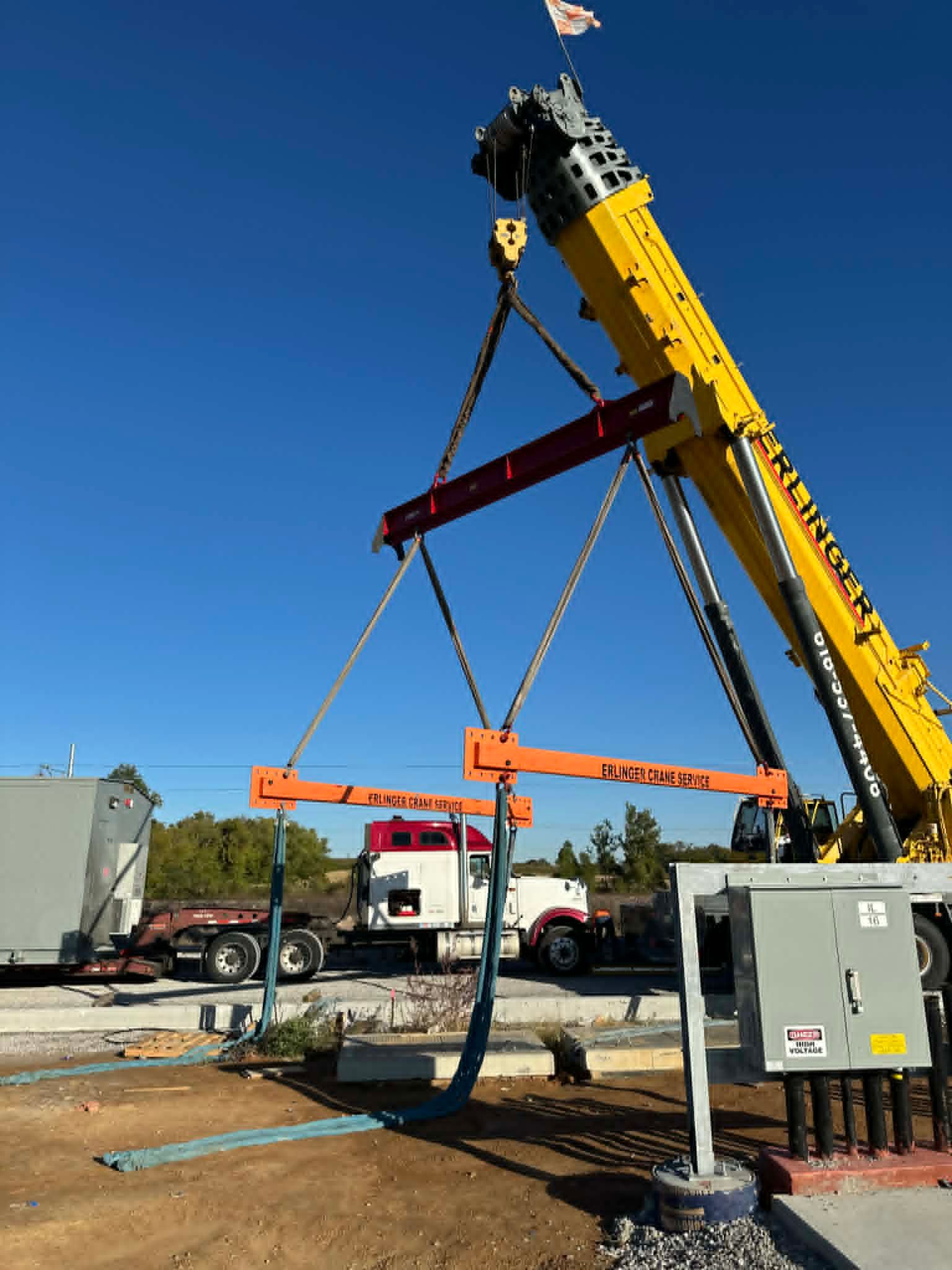 A crane lifts a horizontal steel beam at a construction site on a sunny day.