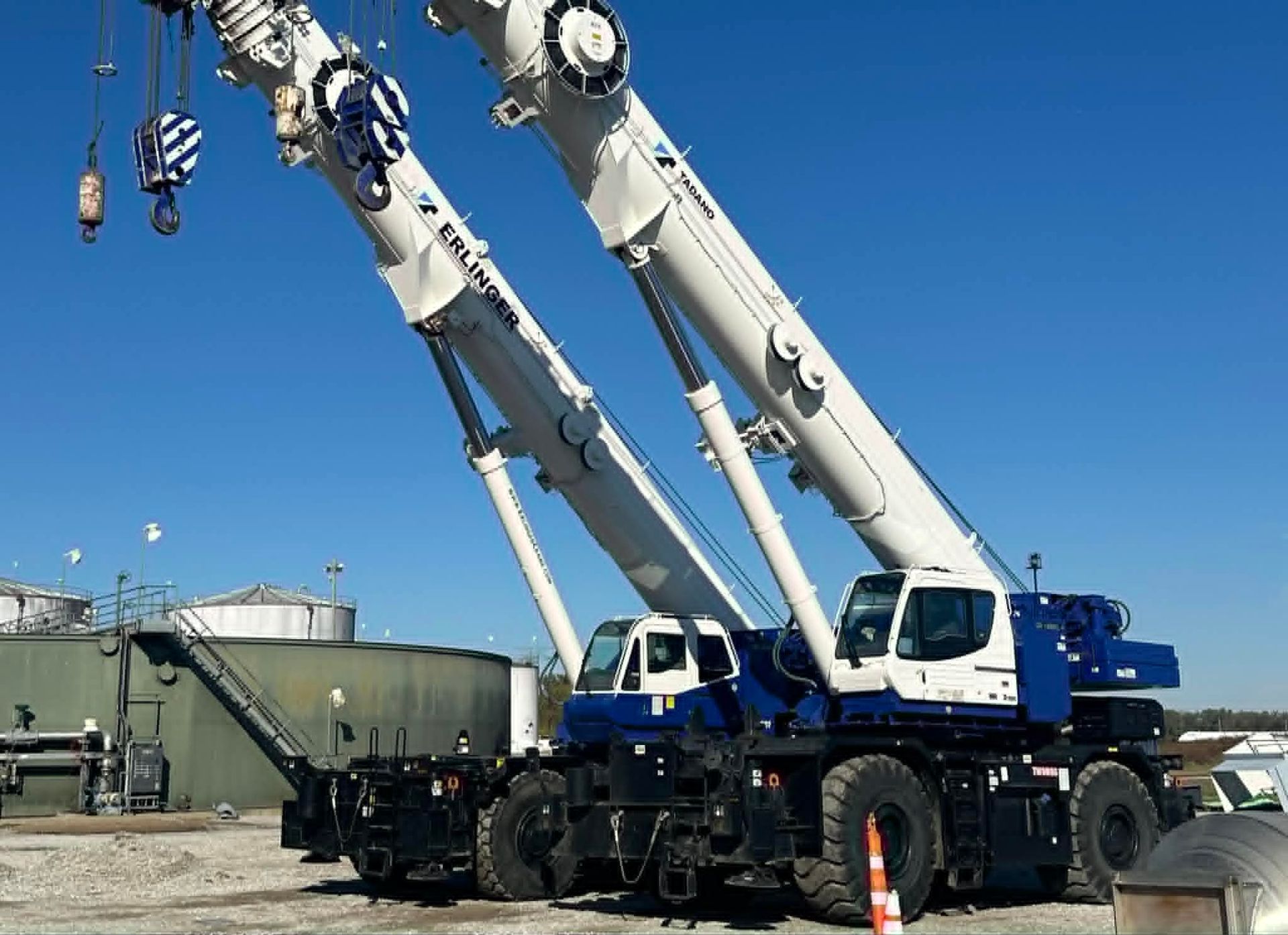 Two white and blue mobile cranes against a clear blue sky, parked near industrial tanks.