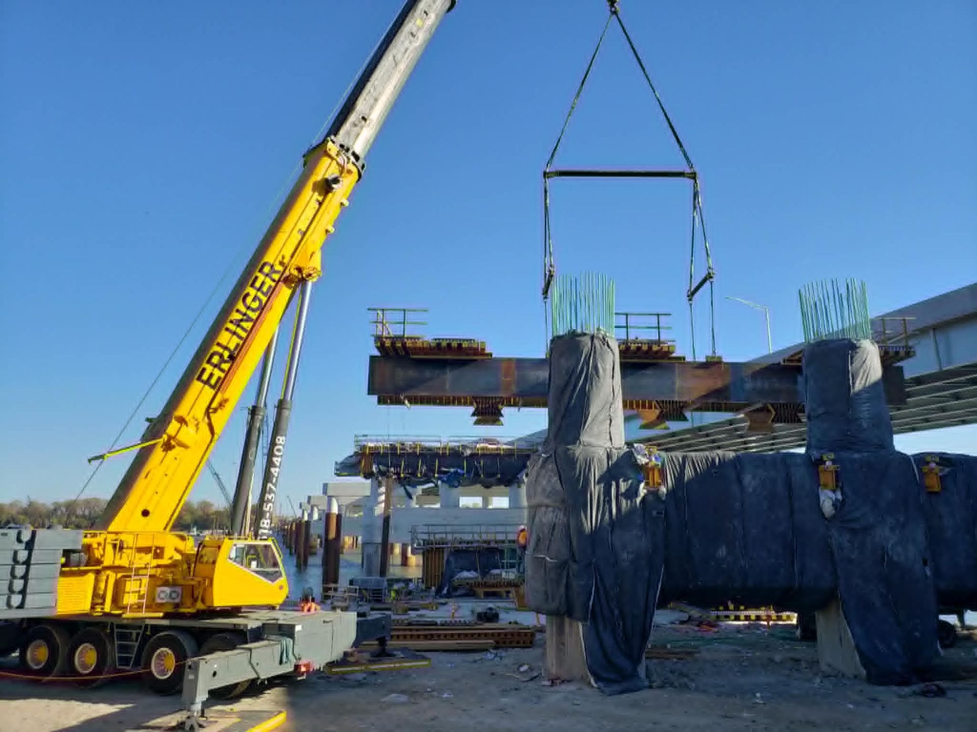 Crane lifting a large steel beam at a bridge construction site; blue sky.