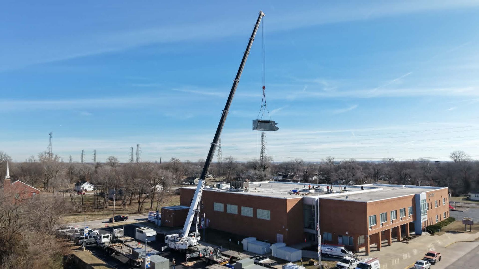 A crane lifting a large HVAC unit onto a brick building on a sunny day.
