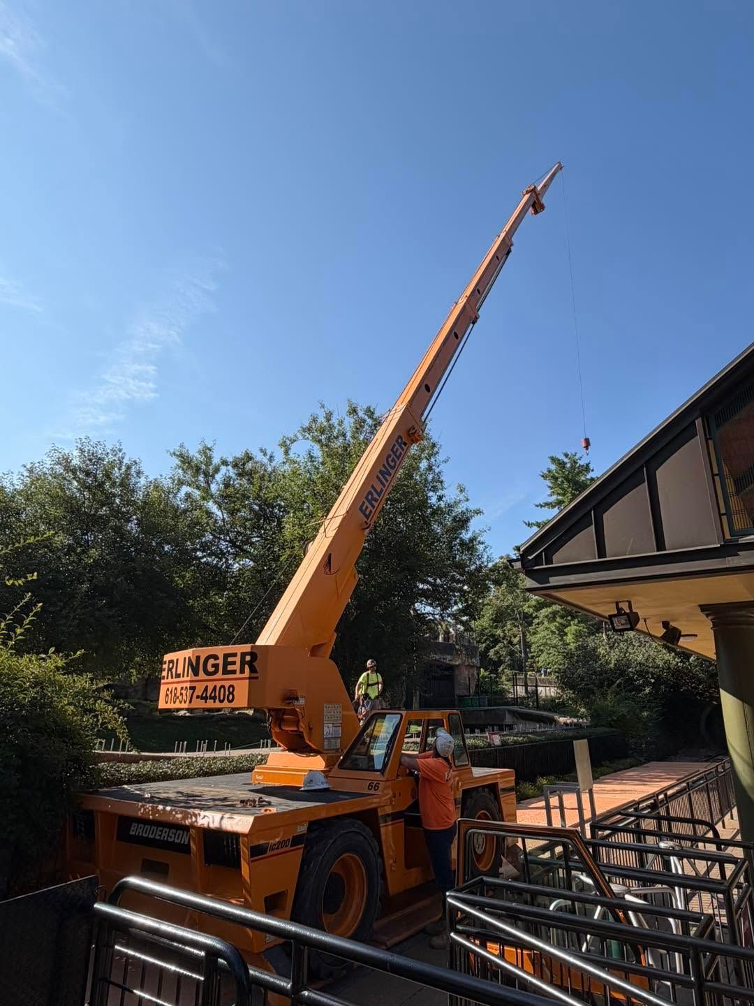 Orange crane lifting a long boom beside a house and metal scaffolding under a blue sky