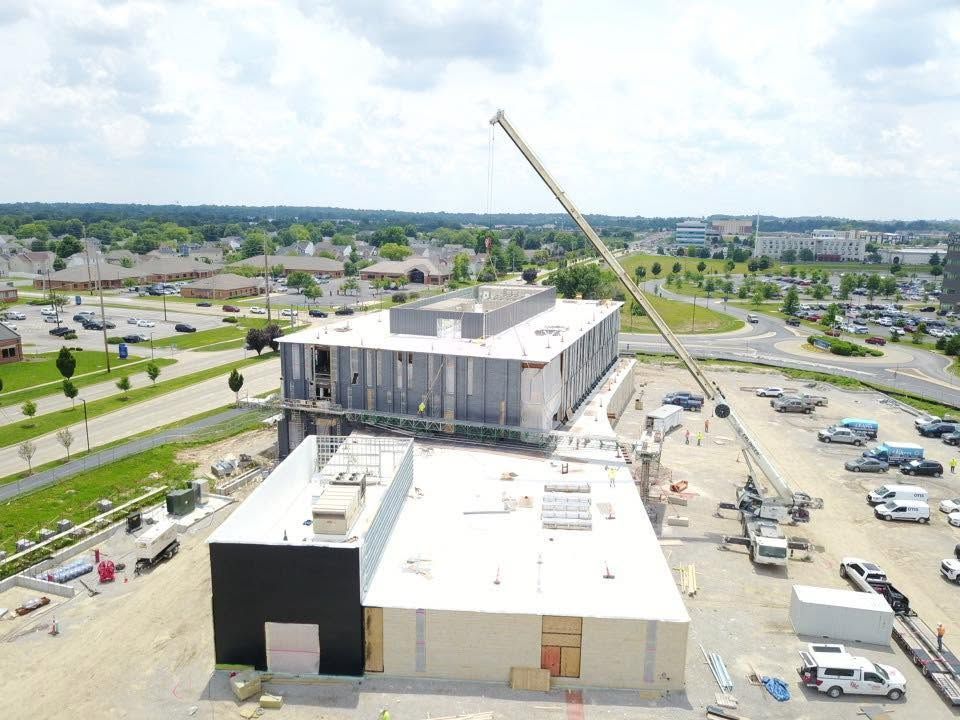 Aerial view of a building under construction with a crane, parking lots, and nearby roads under a bright sky.
