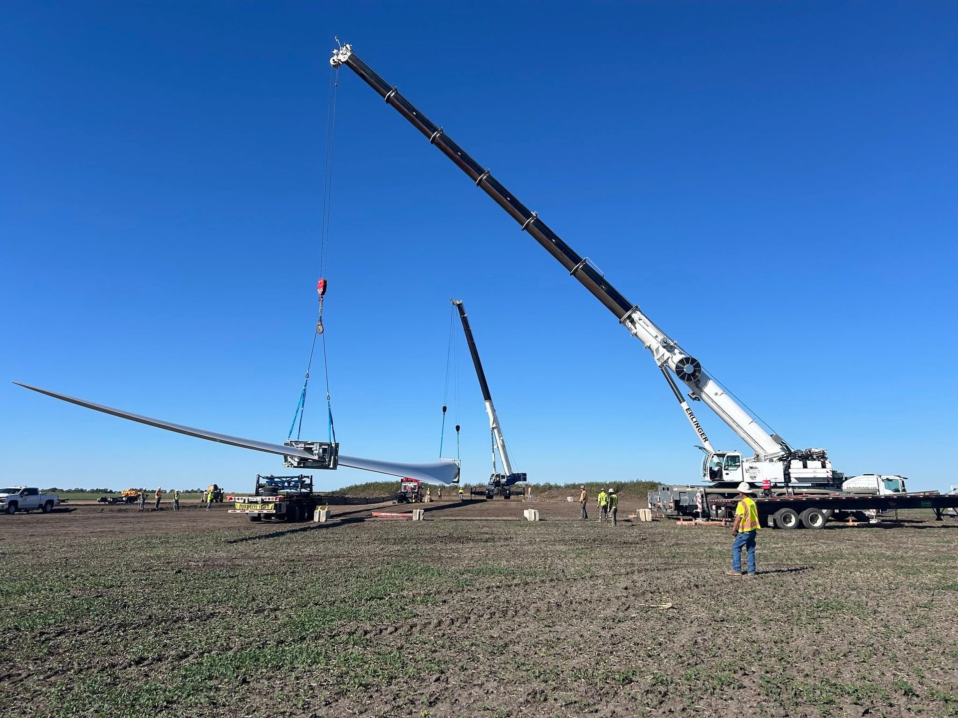 Crane crew lifting a long wind turbine blade in a field under a blue sky