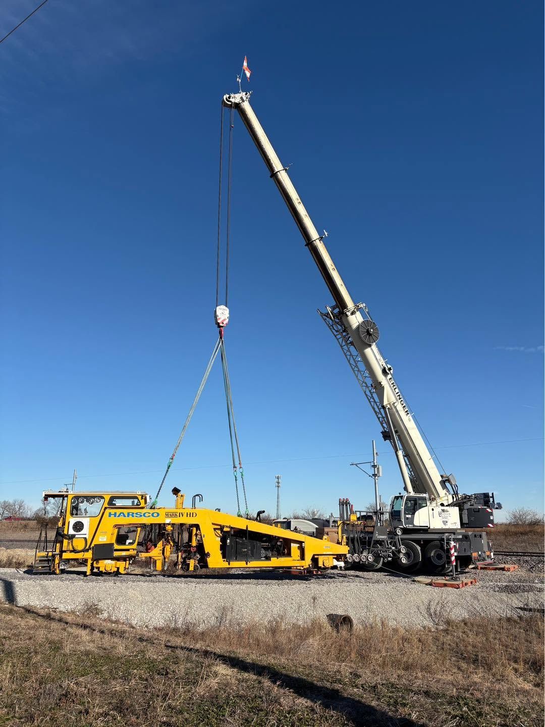 Yellow crane lifting a long yellow industrial machine in a gravel lot under a clear blue sky
