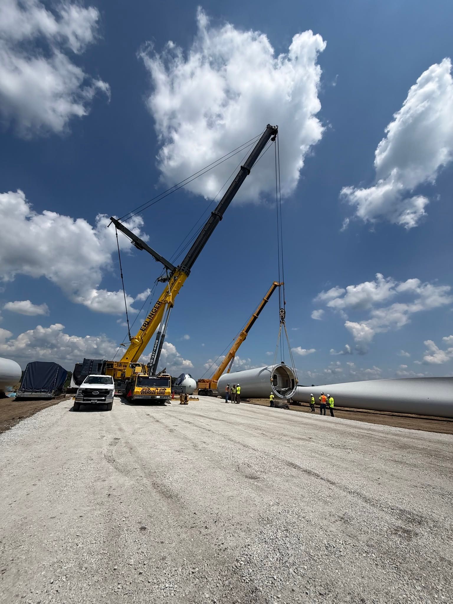 Construction cranes lifting a large white wind turbine blade on a gravel site under a cloudy blue sky