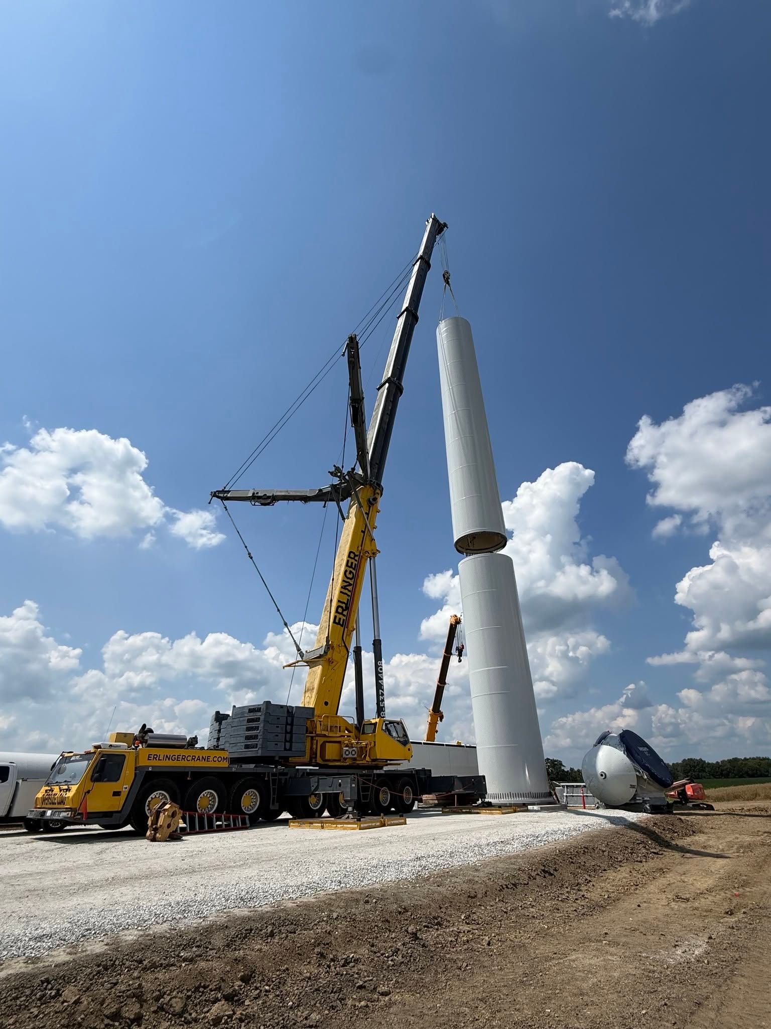 Wind turbine tower being installed by a yellow crane on a gravel site under a blue sky.