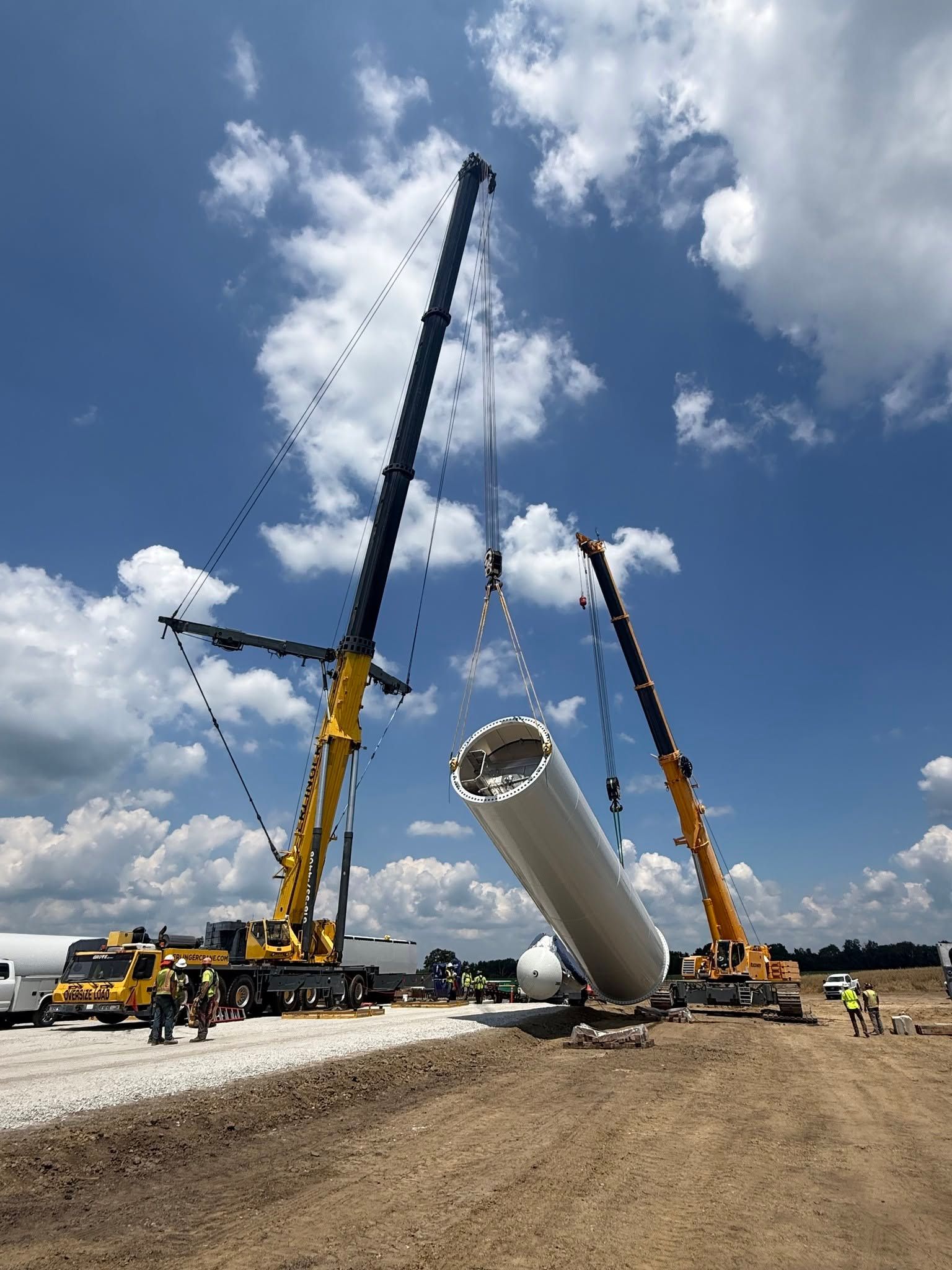 Construction cranes lifting a large white cylindrical structure under a blue sky with clouds