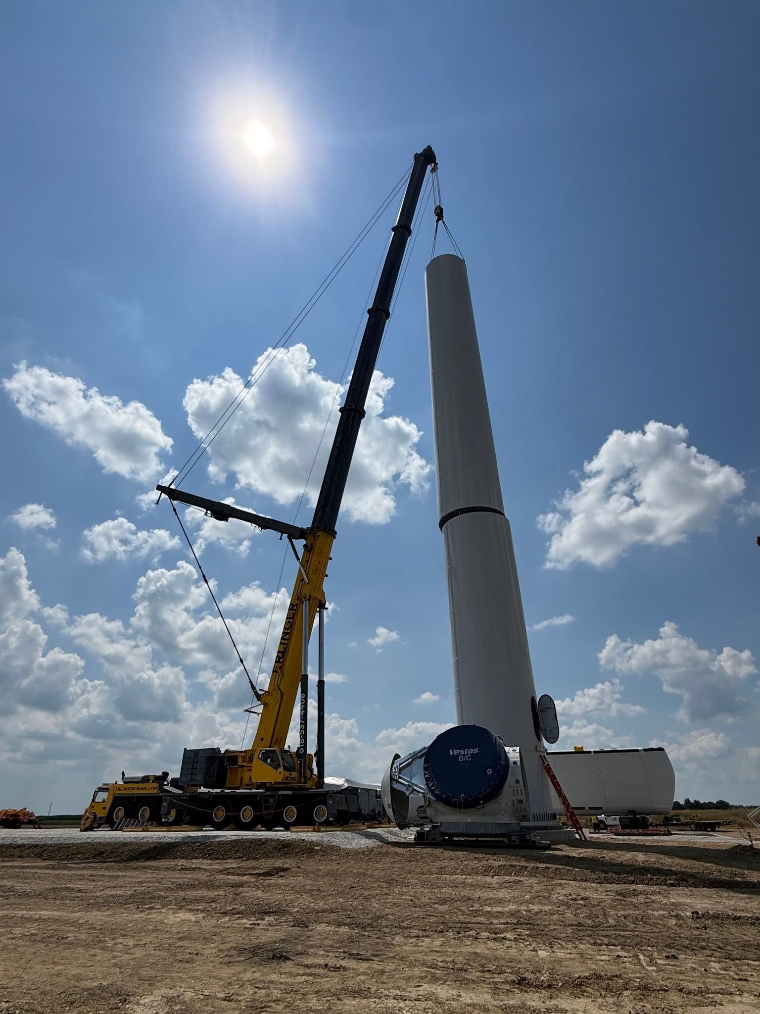 Crane lifting a large white wind turbine tower section at a construction site under a bright blue sky.