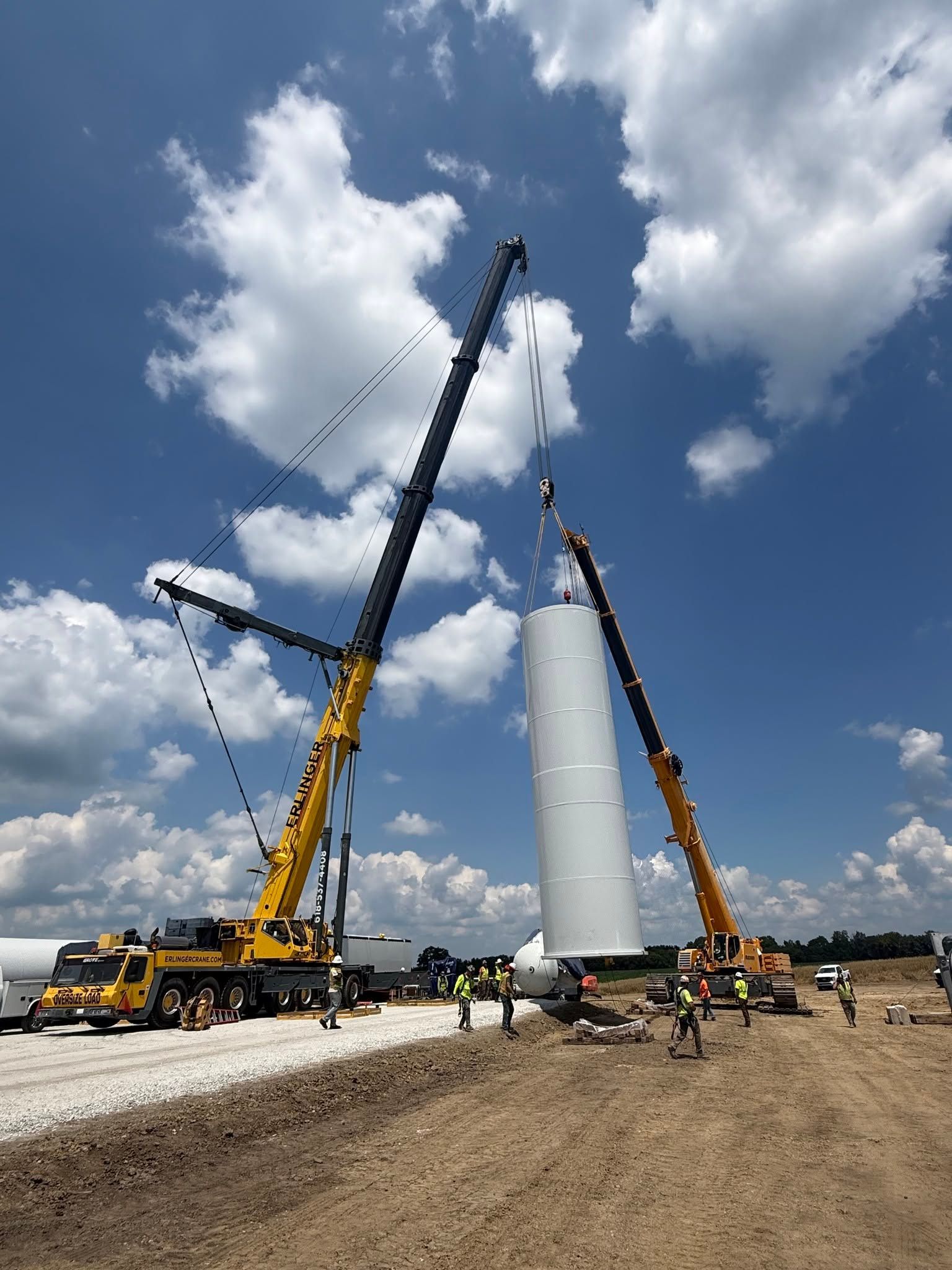 Crane crews lifting a tall white wind turbine tower section on a construction site under a blue, cloudy sky