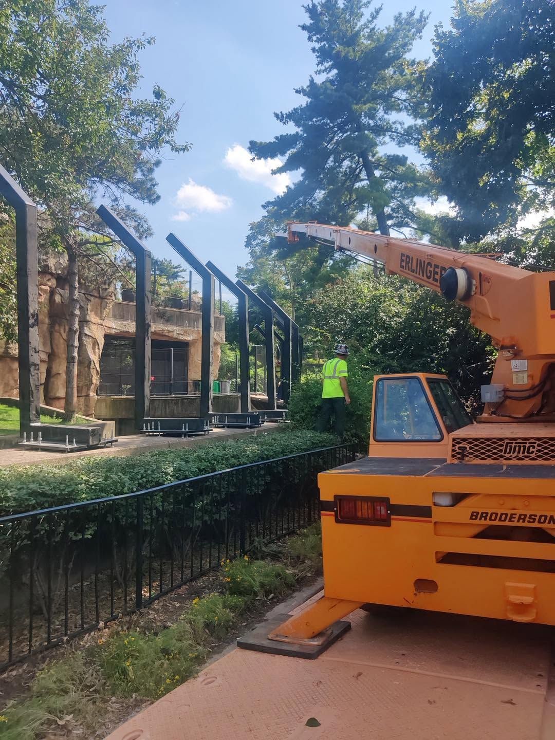 Orange construction vehicle beside a tree-lined path near a brick building under renovation