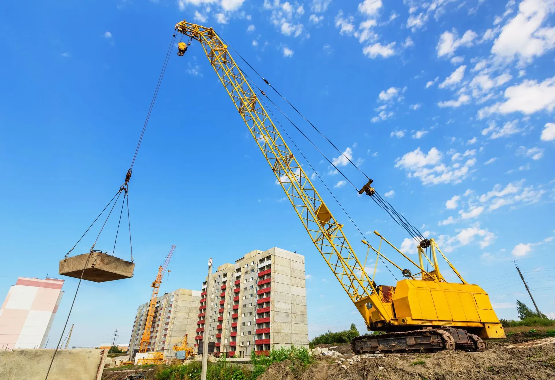 Yellow construction crane lifting a large concrete block at a construction site with apartment buildings under a blue sky.