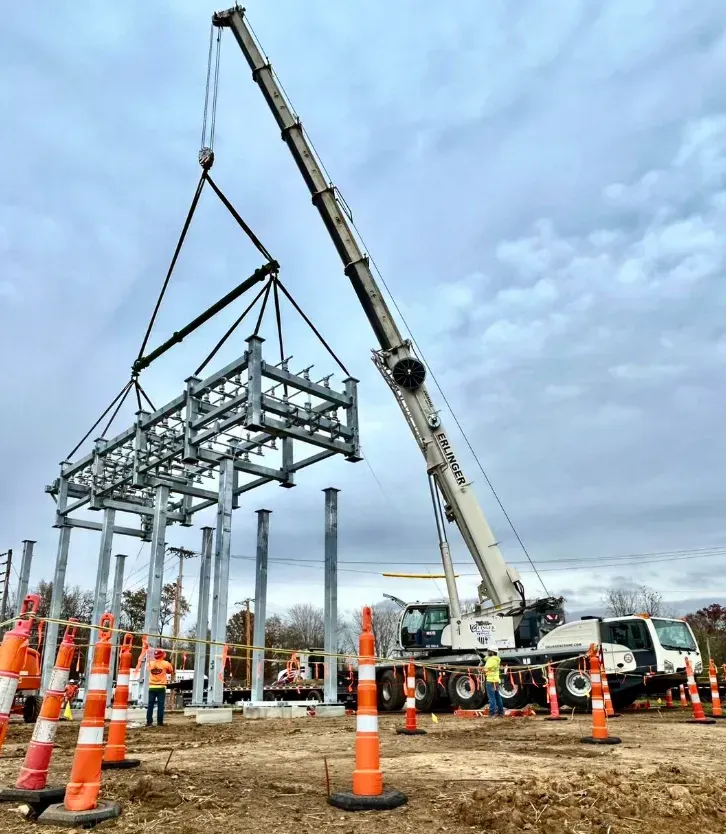 Crane lifting electrical grid component onto support pillars at construction site. Workers and traffic cones present.