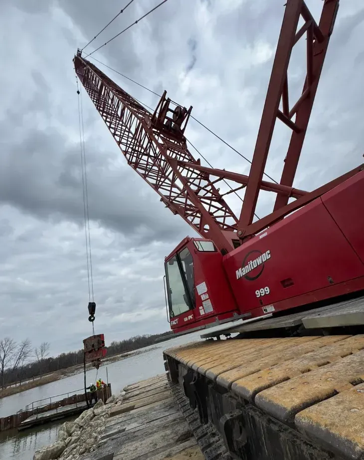 Red crane with boom over water, lifting a load. Overcast sky.
