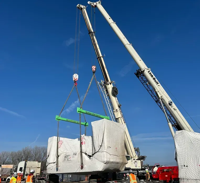 Two cranes lifting a large, wrapped block; workers nearby in bright clothing under a blue sky.