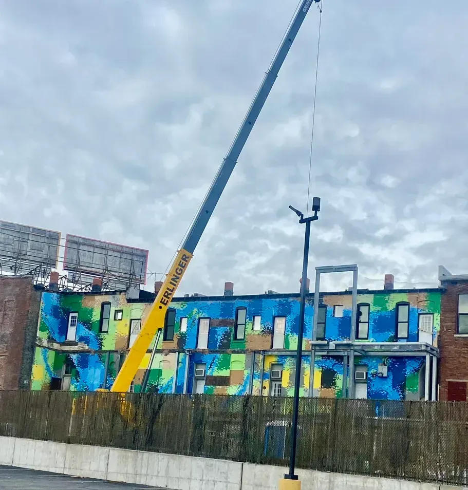 A tall yellow crane extends toward a colorful mural on a building under a cloudy sky.