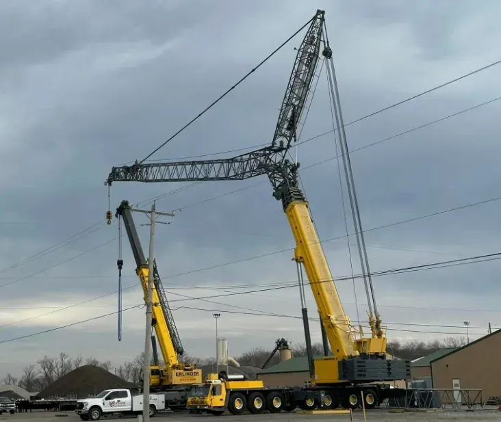 Two yellow cranes near power lines under a cloudy sky. One crane is lifting a utility pole.