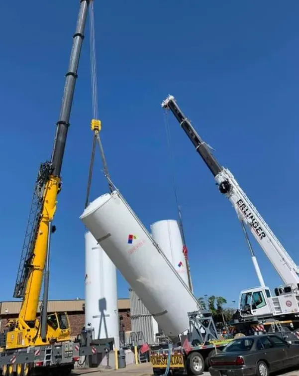 Two cranes lifting large white cylindrical tanks outdoors against a blue sky.