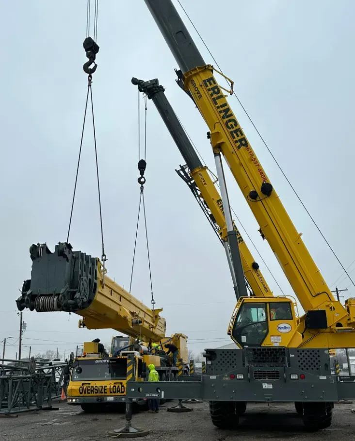 Two yellow cranes lifting a large, yellow machinery piece in an outdoor setting.