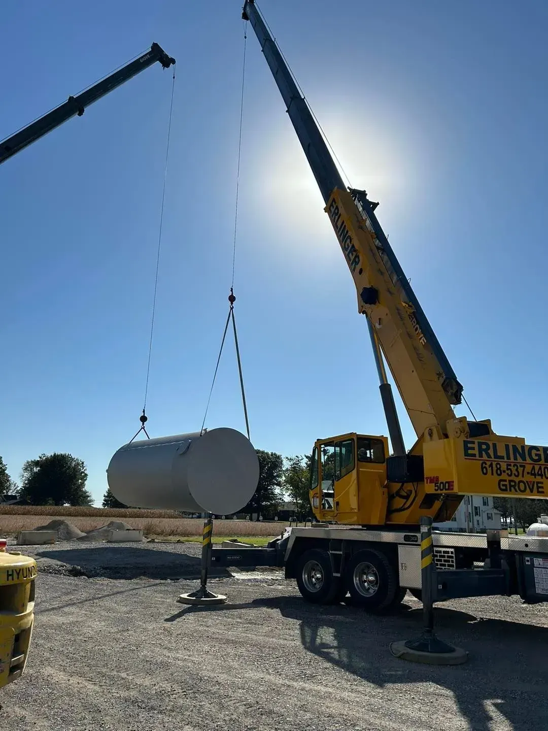 Two cranes lifting a large cylindrical tank on a construction site under a bright sun.
