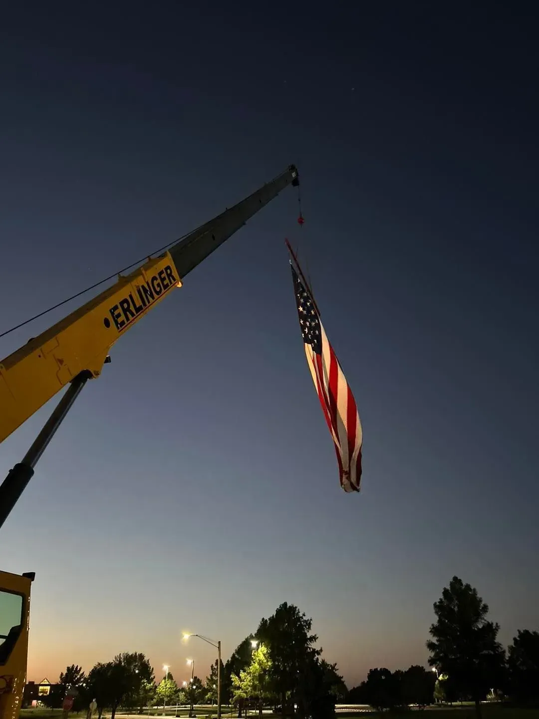 American flag suspended by a yellow crane against a dusk sky.