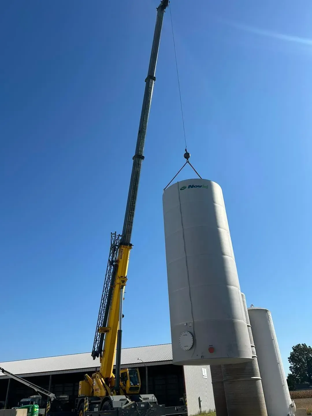 A tall crane lifting a cylindrical tank against a blue sky. Another similar tank is nearby.