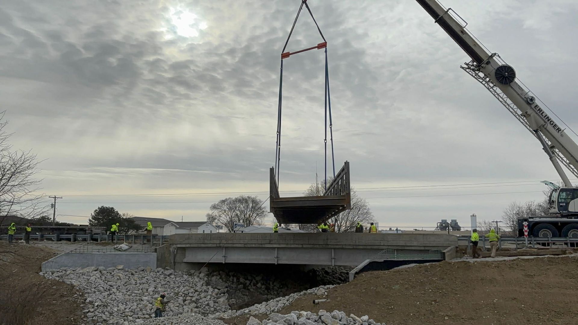 Crane lifting a bridge section over a roadway; construction workers in safety vests; cloudy sky.