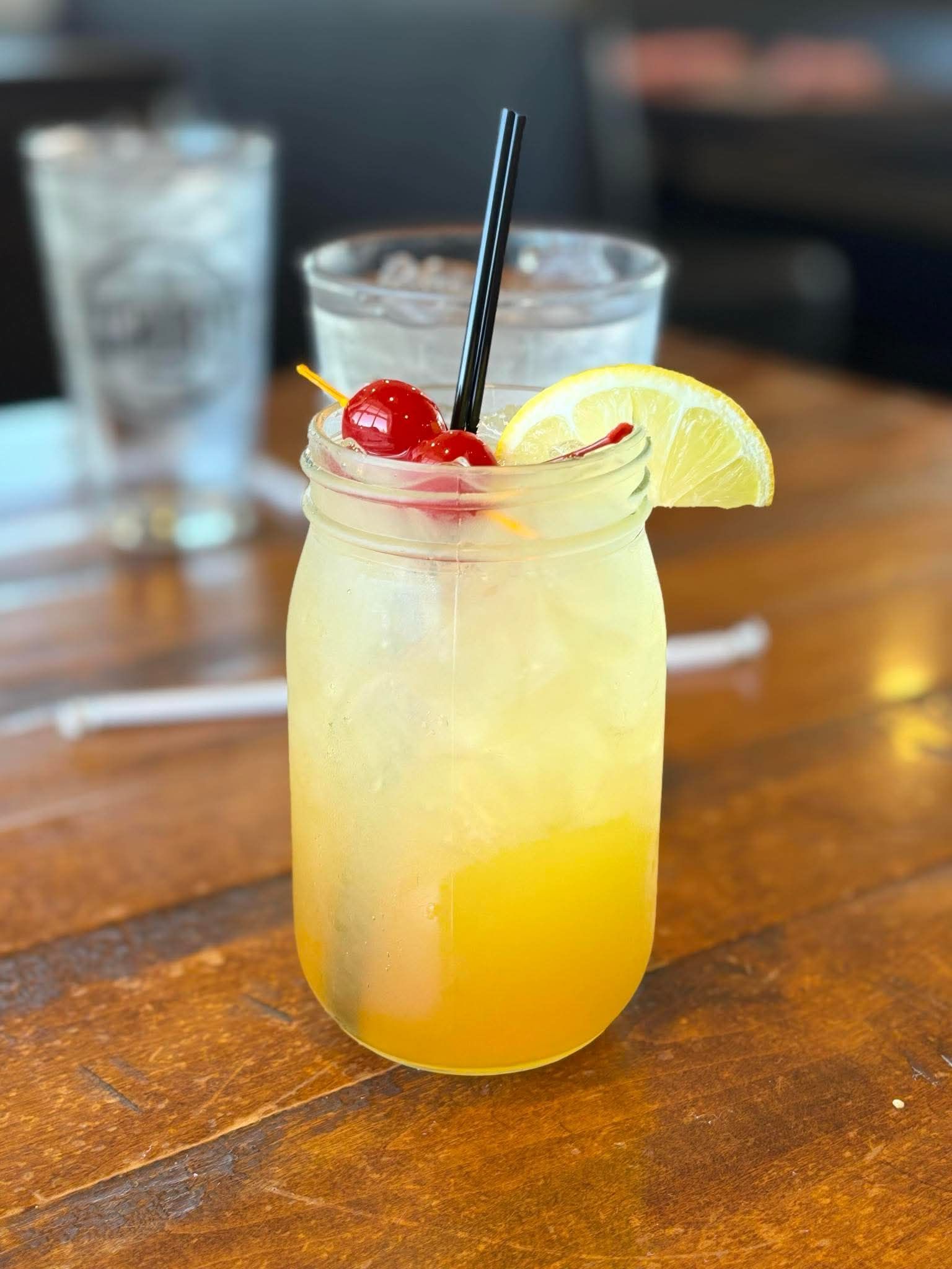 A tropical cocktail in a mason jar, garnished with cherries and a lemon slice, on a wooden table.