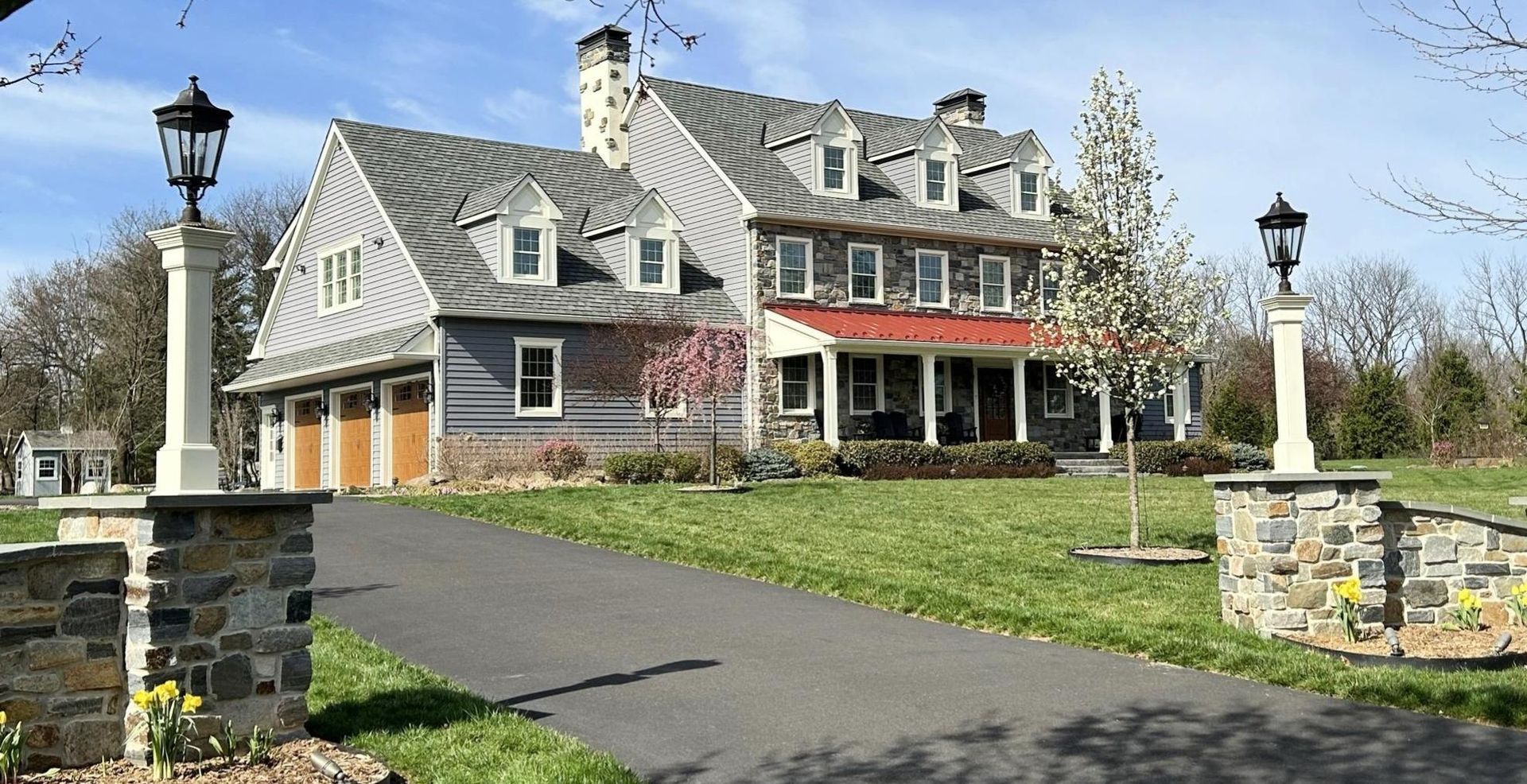 Two-story house with stone pillars, a driveway, and two lamp posts. The house has a gray roof and siding.