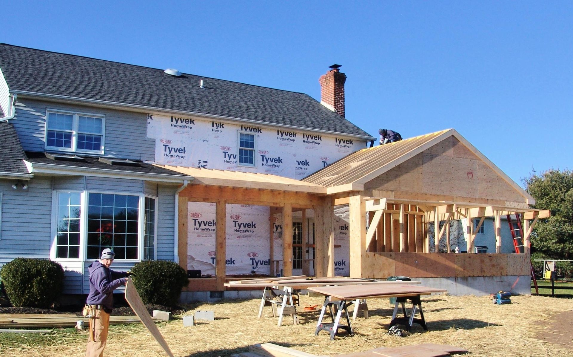 Construction of a wood-framed porch addition to a two-story house; workers are present, Tyvek wrap visible.