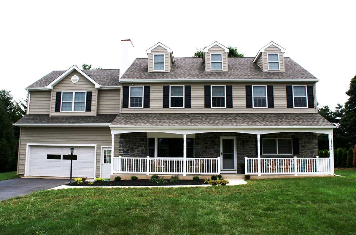 Two-story beige house with a stone porch, white railing, and a garage. Black shutters and three dormer windows.