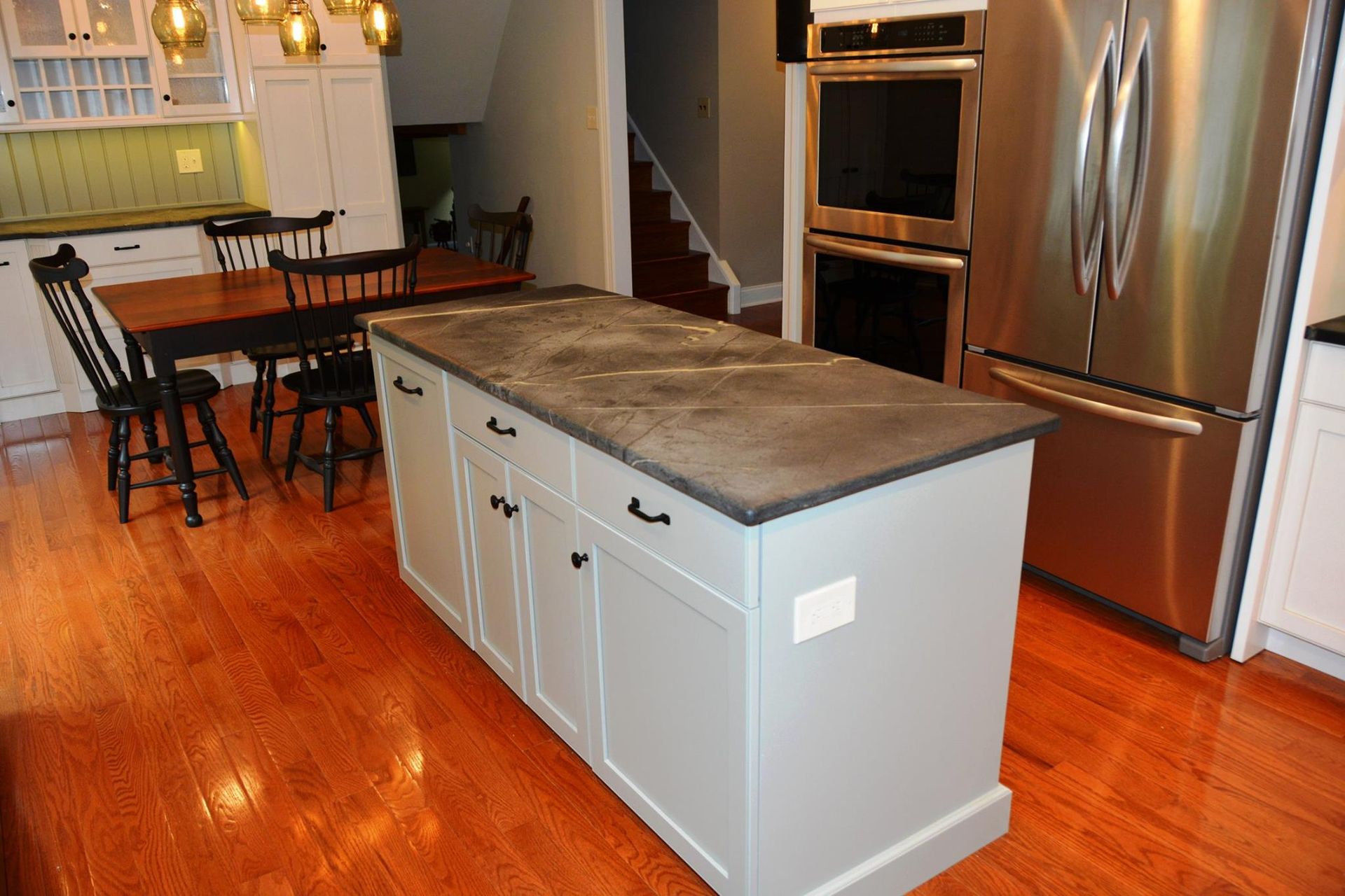 Kitchen with gray island and countertop, stainless steel refrigerator, wooden table, and hardwood floor.