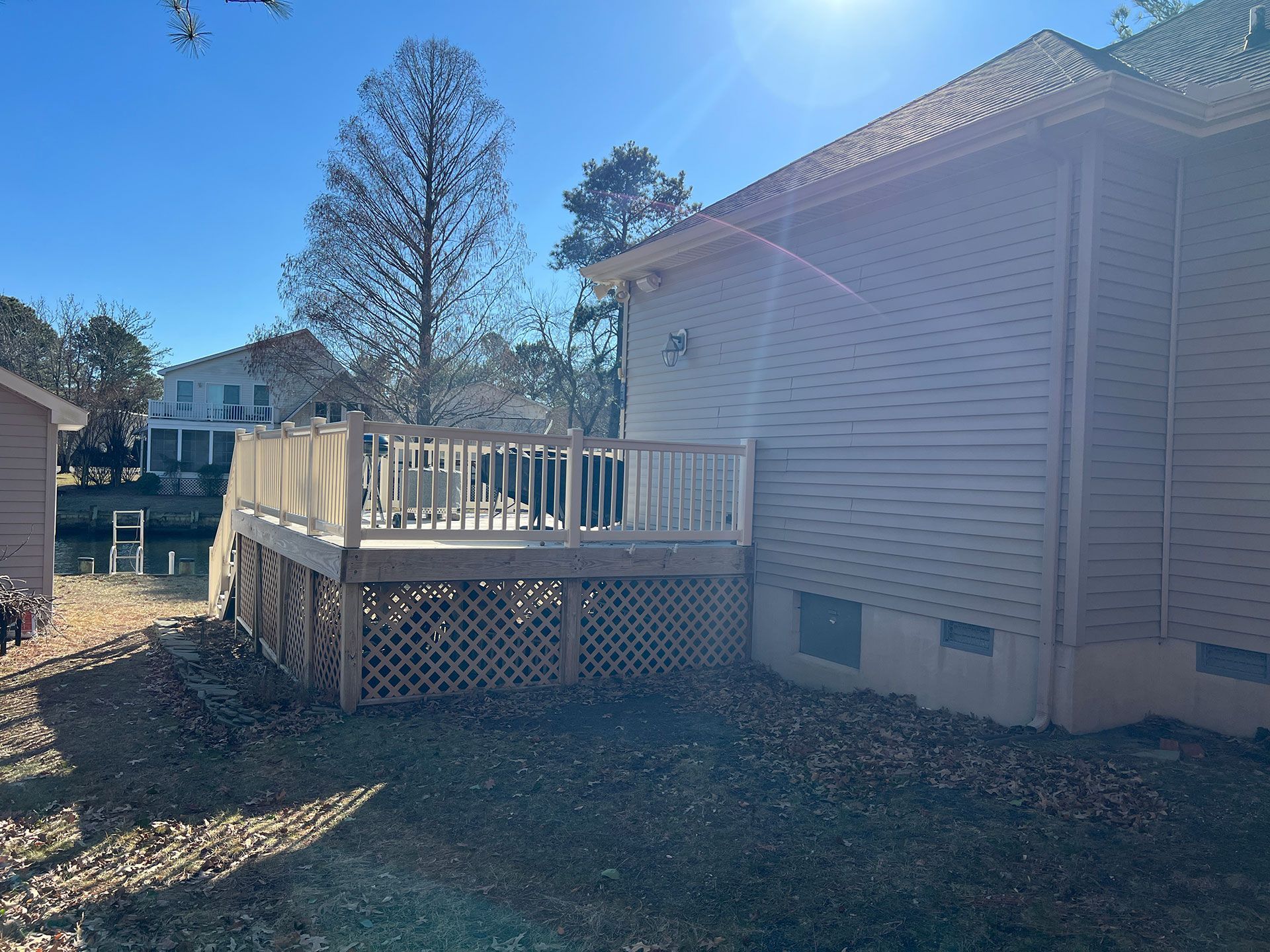 Wooden deck next to a house with a latticework base, overlooking a waterway on a sunny day.