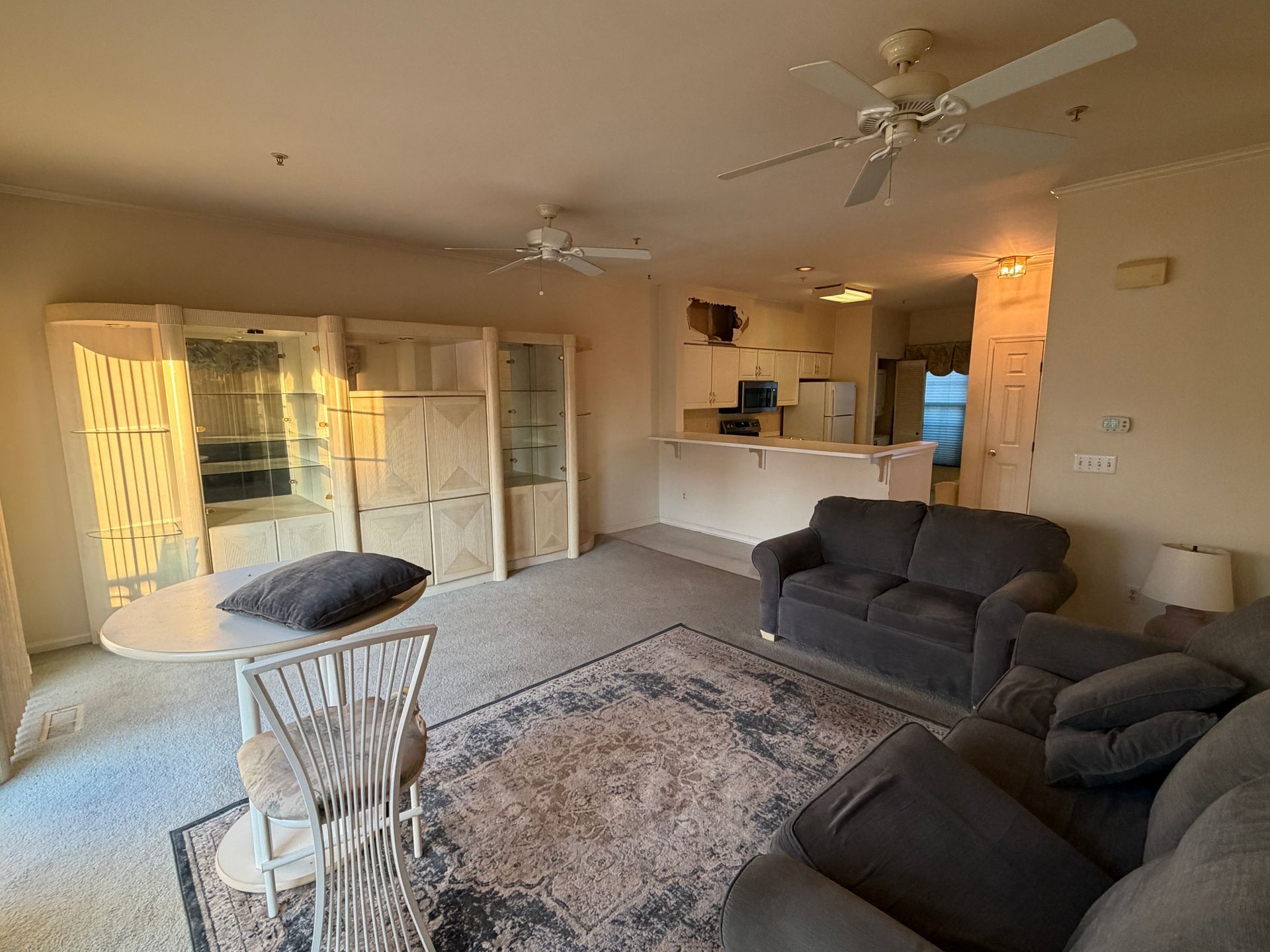 Bright living room with gray sofas, white rug, dining chairs, and open kitchen in the background