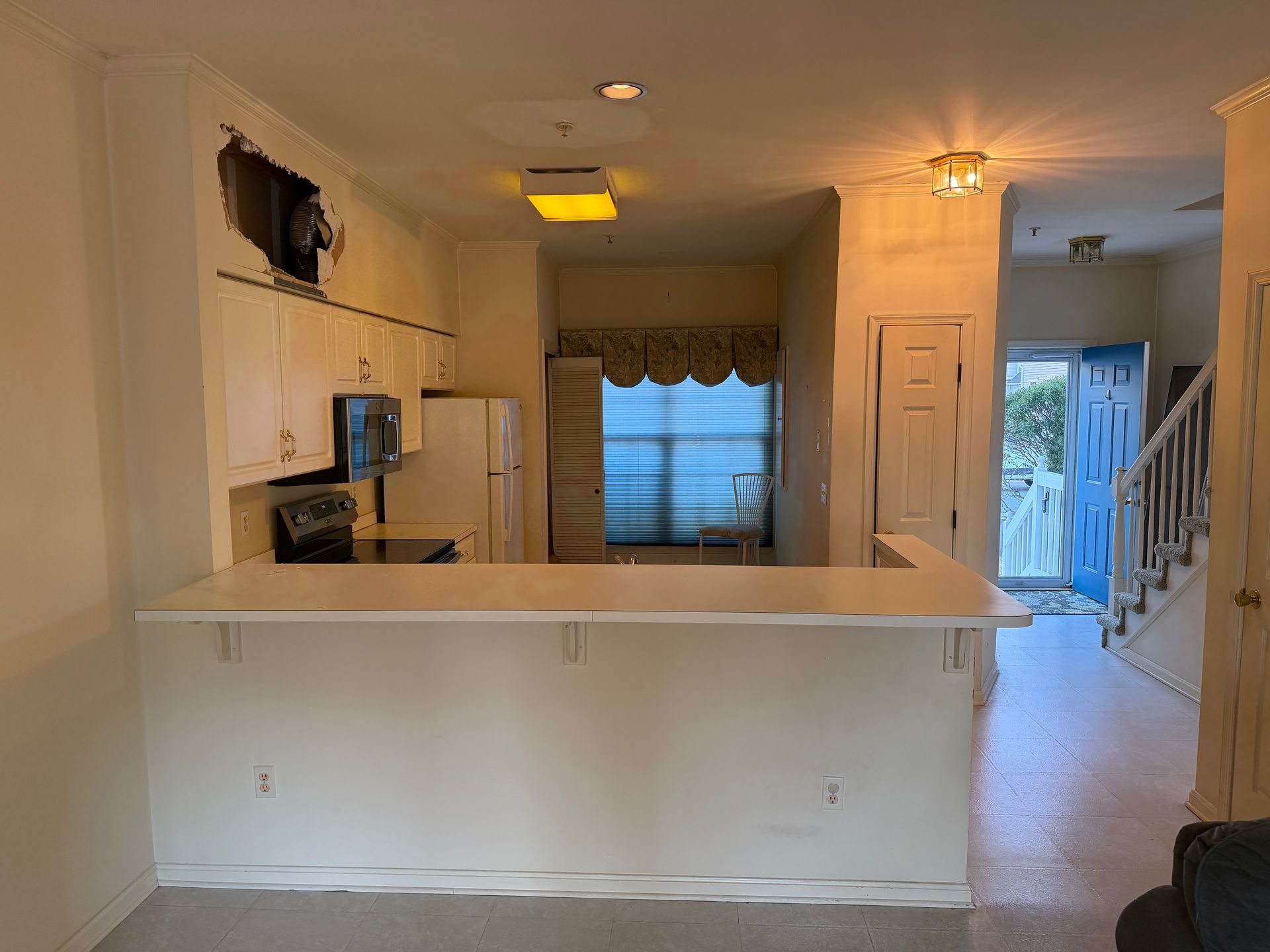 Empty white kitchen and dining area with a pass-through counter, window curtains, and hallway beyond