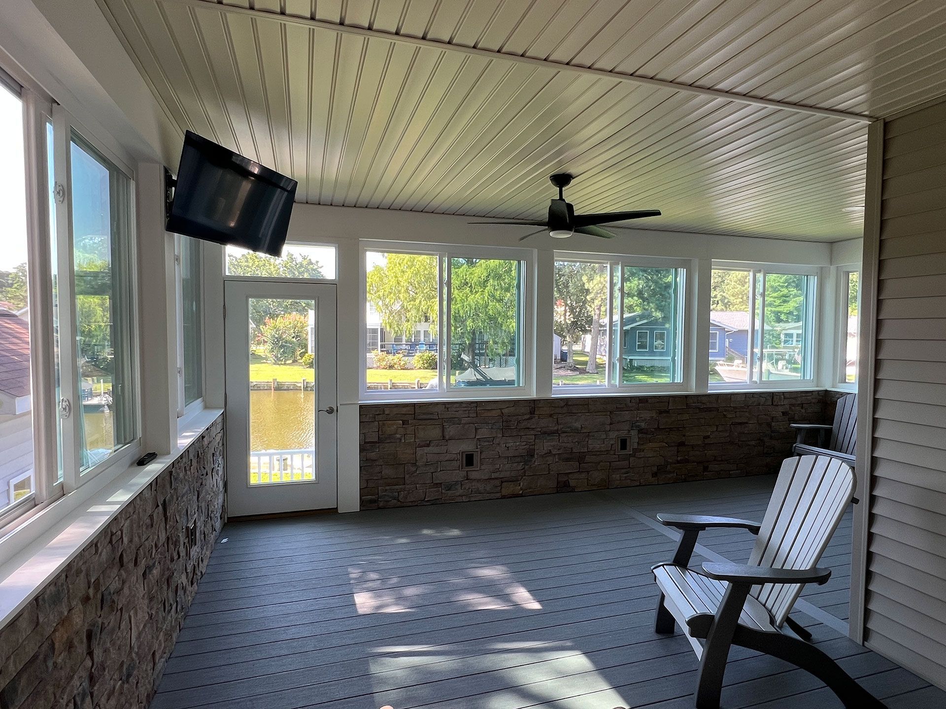 Sunroom with windows, brick accent wall, ceiling fan, television, and an Adirondack chair.