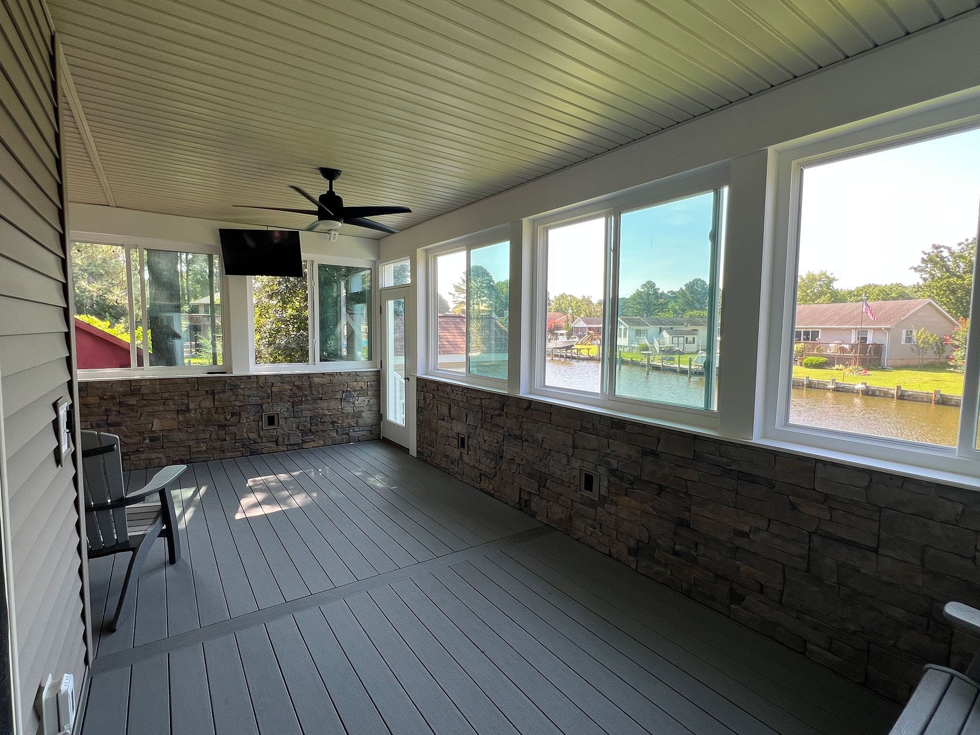 Covered porch with stone accent wall, gray deck, windows overlooking water.