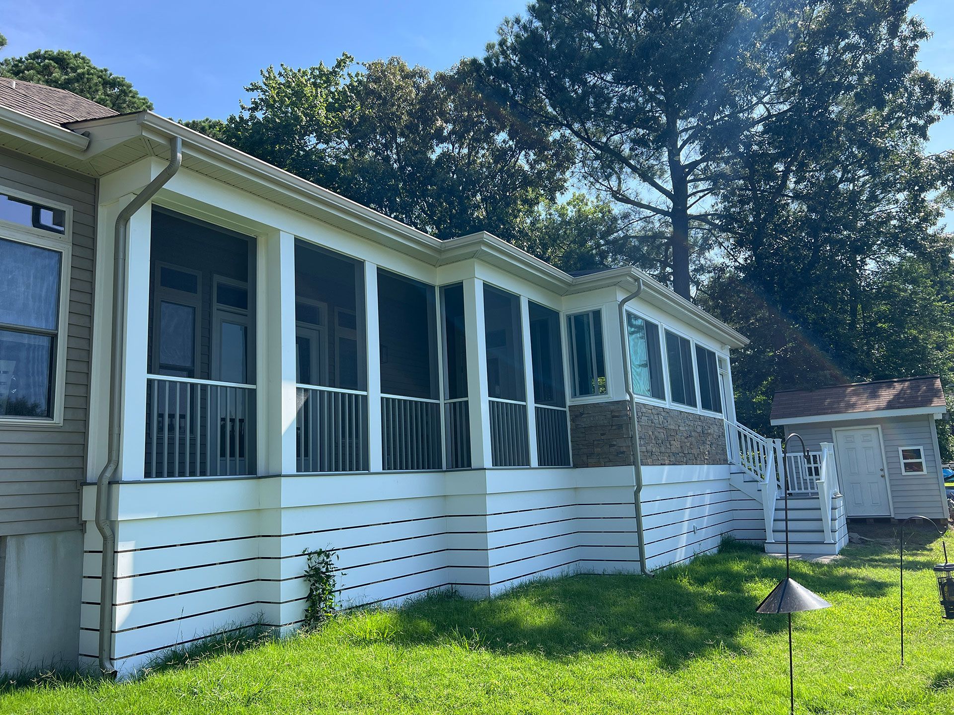 A screened porch with white trim and railings on a green lawn, near a small building, sunny day.