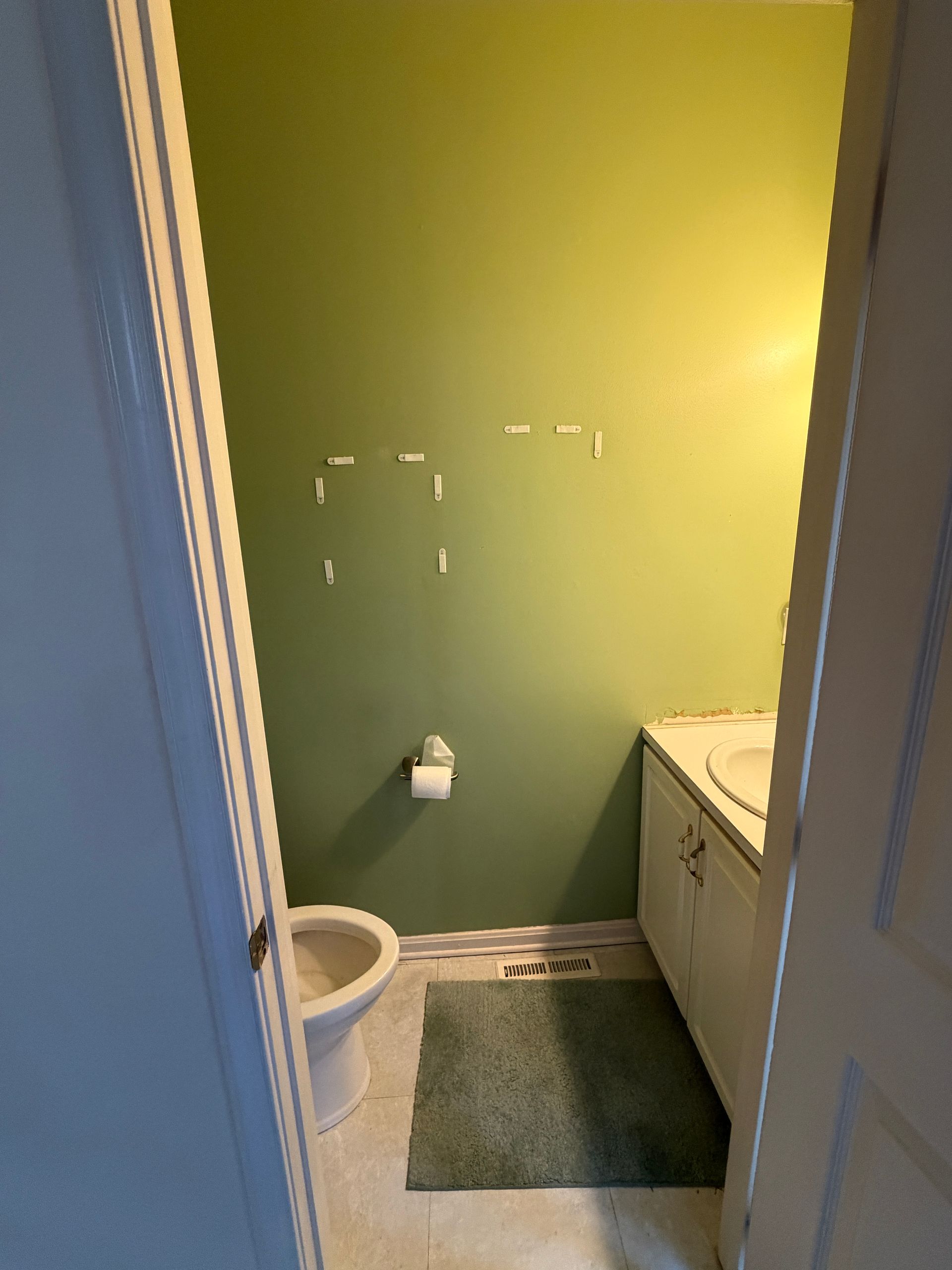 Small bathroom with green walls, toilet, towel holder, sink cabinet, and a bath mat.