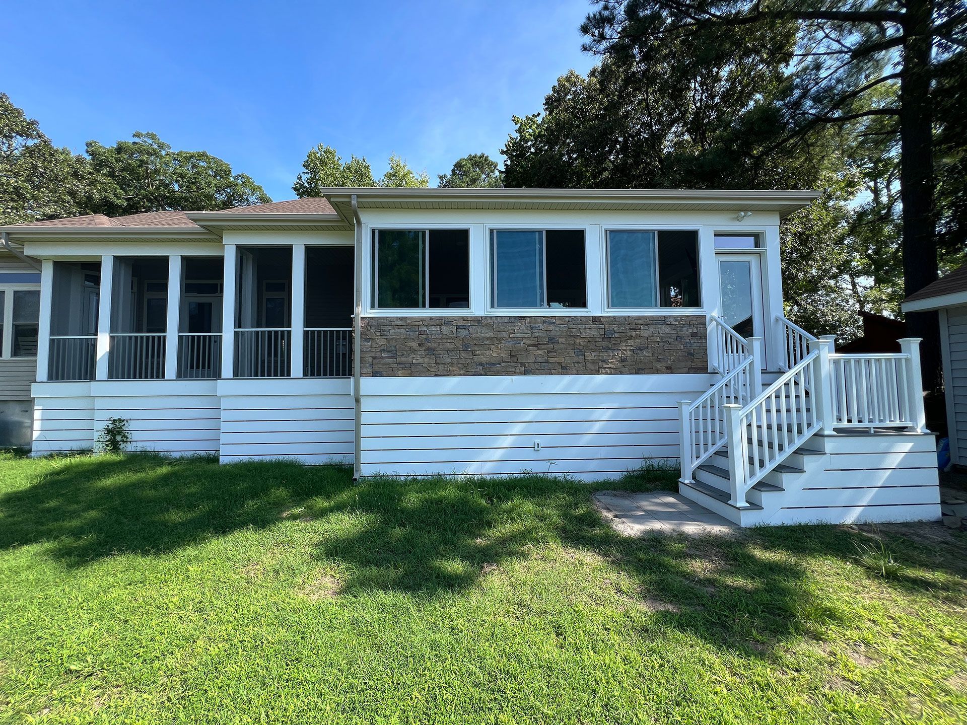 White cottage with screened porch, windows, and steps on a grassy lawn under a blue sky.