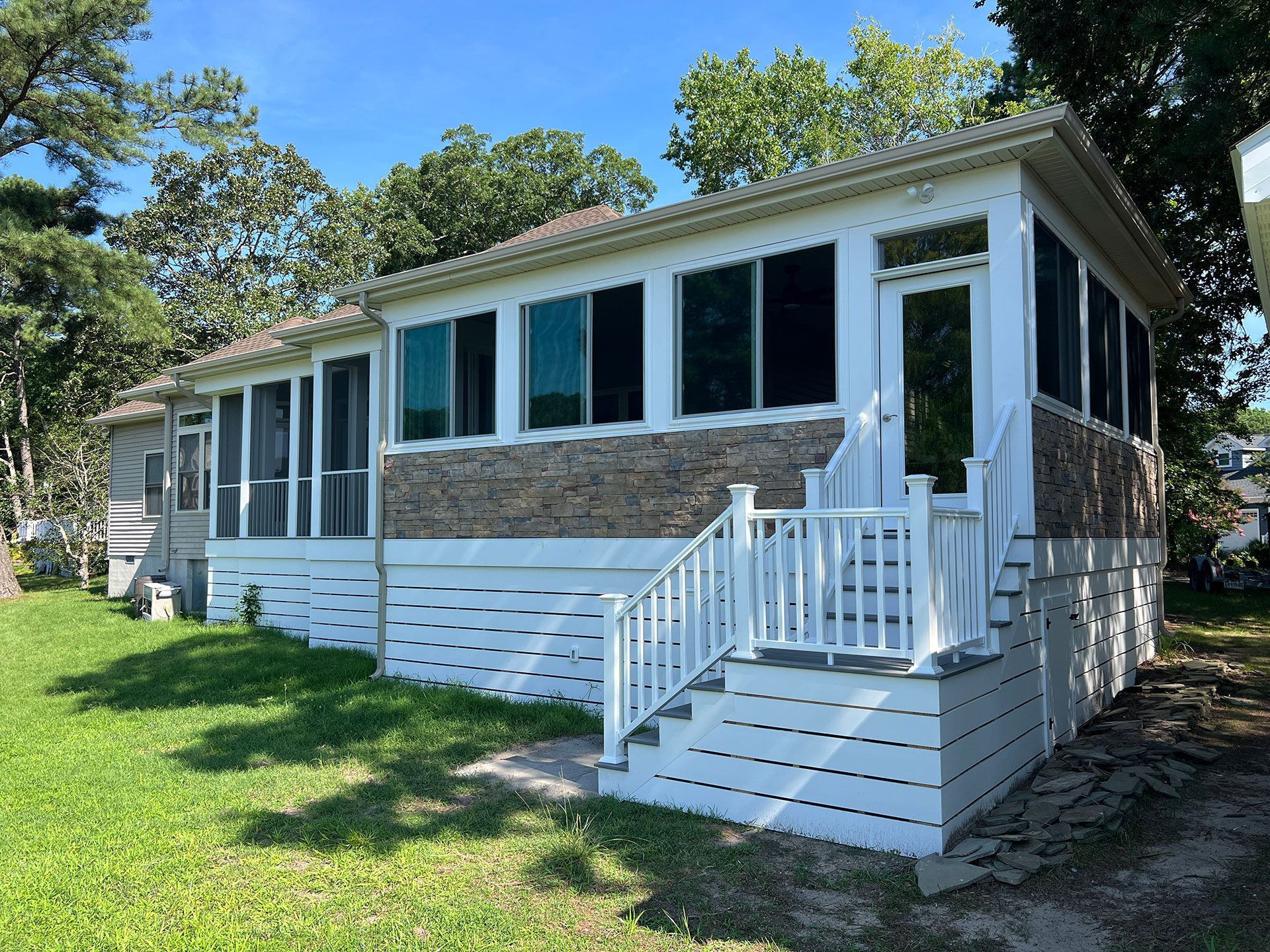White porch with steps, screens, and windows on a house with wood siding. Green grass, trees.