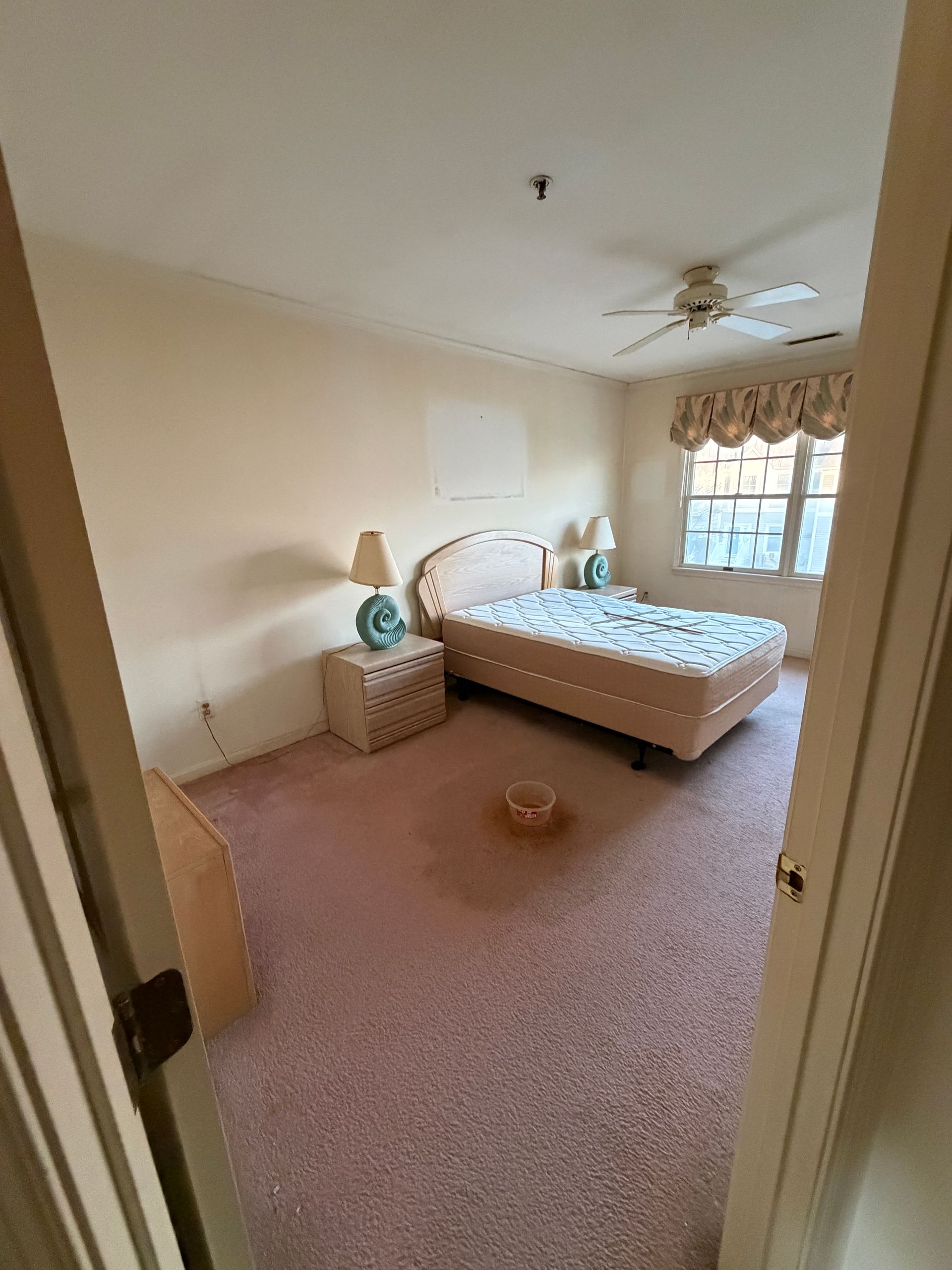 Small beige bedroom with a bed, nightstand, ceiling fan, and window curtains visible through an open doorway