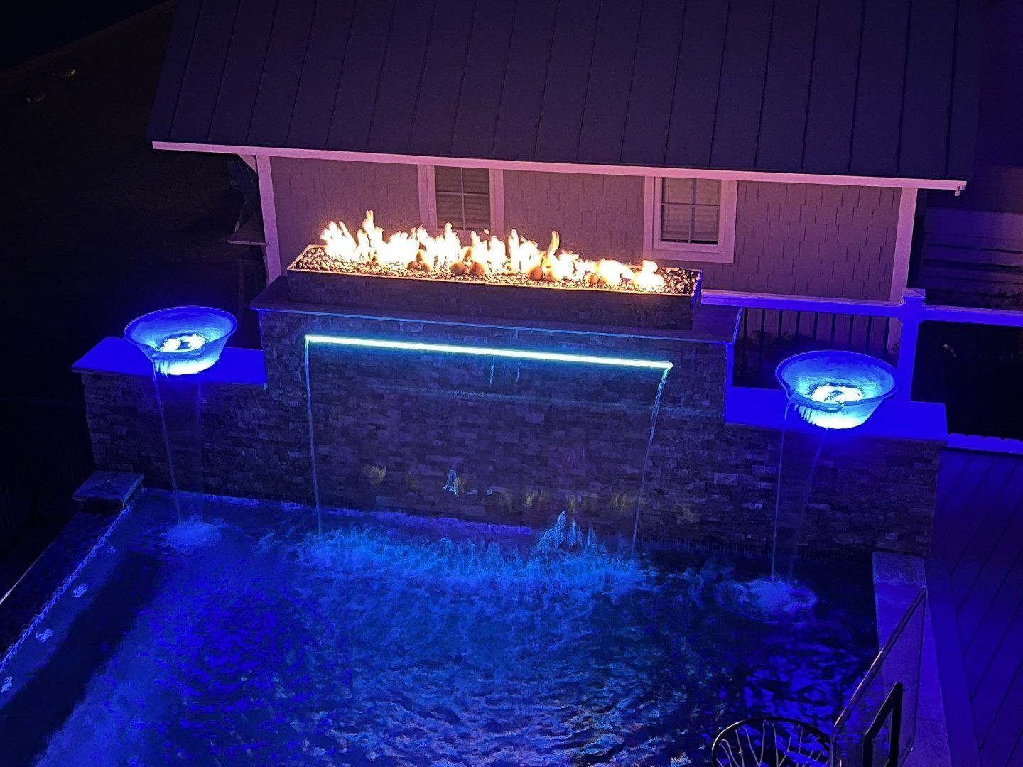 A swimming pool with a fire pit and a waterfall at night.