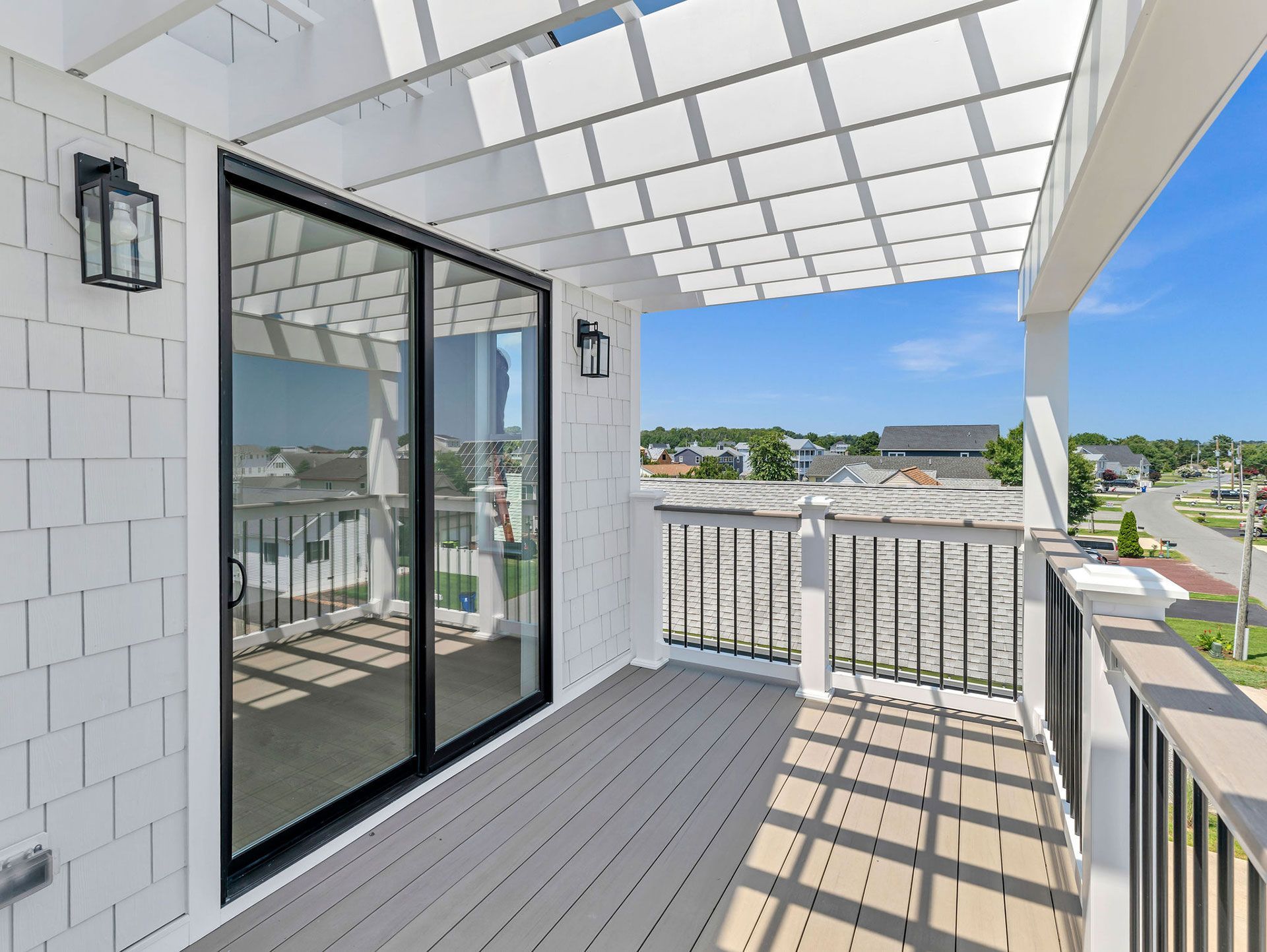 Balcony with pergola, sliding glass door, white siding, gray deck, and black railing overlooking town.