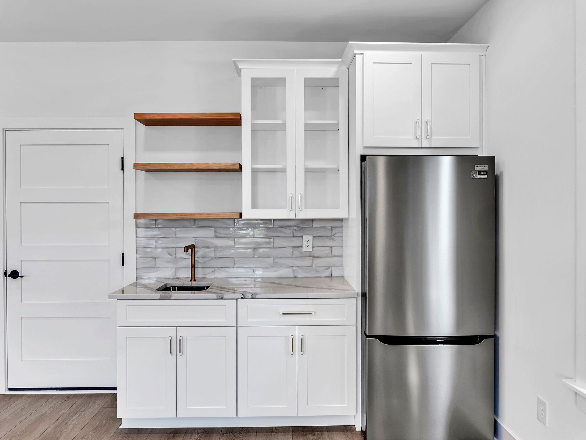 Small white kitchenette with stainless steel refrigerator. Includes sink, cabinets, shelves, and a door.