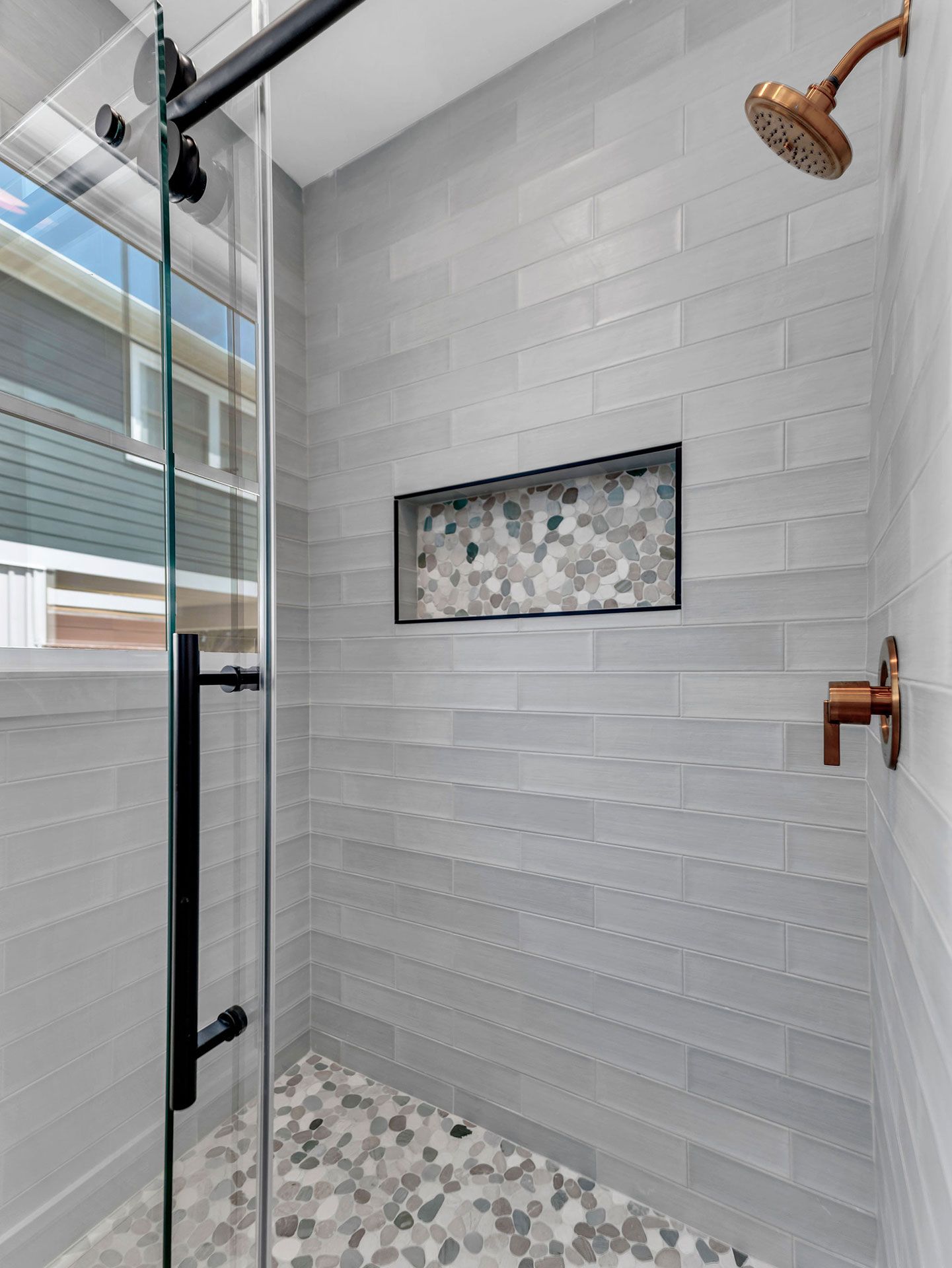 Gray tiled shower with a glass door, pebble floor, and bronze fixtures.
