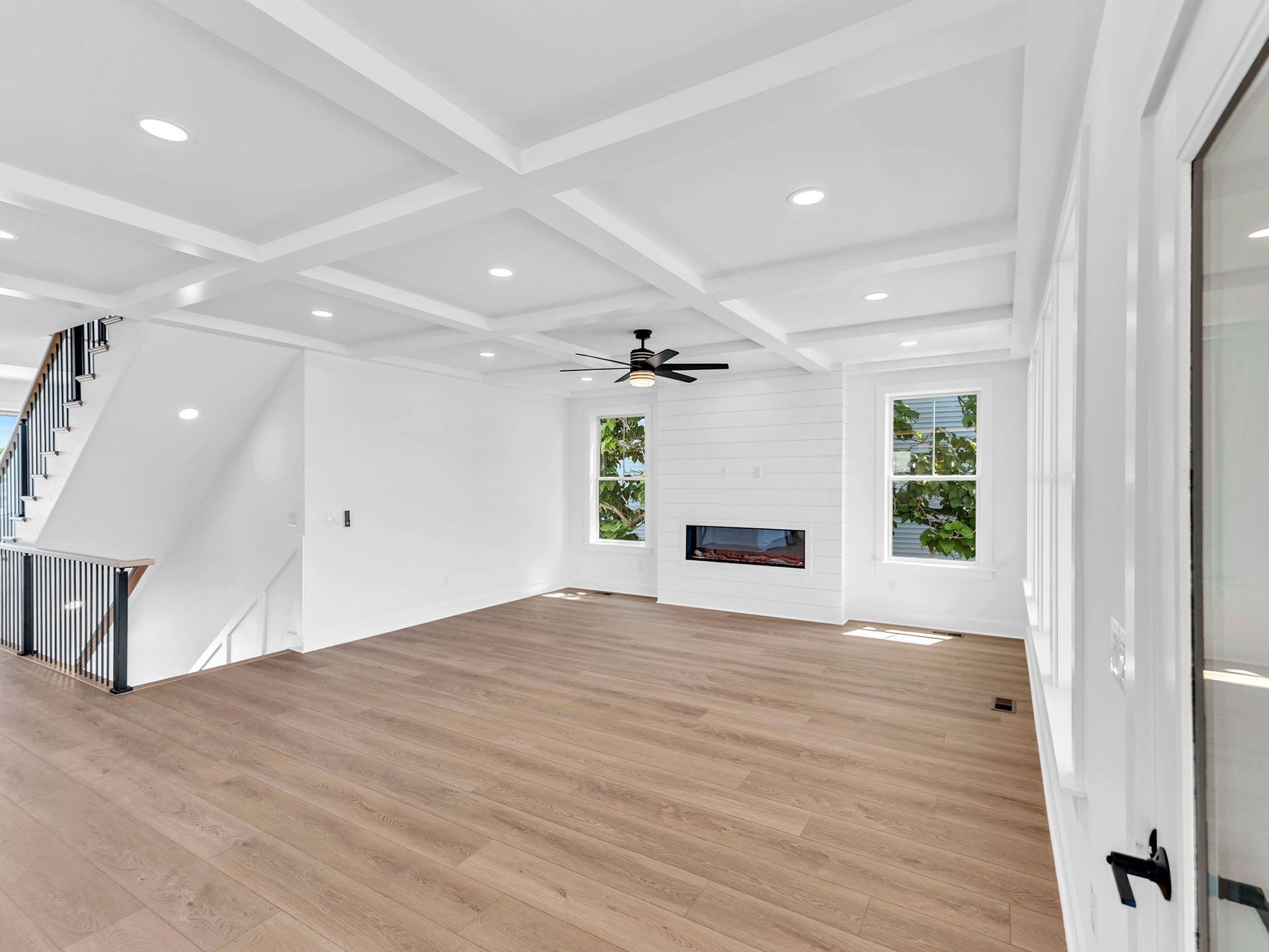 Empty living room with wood floor, white walls and coffered ceiling, fireplace, windows, and stairs.