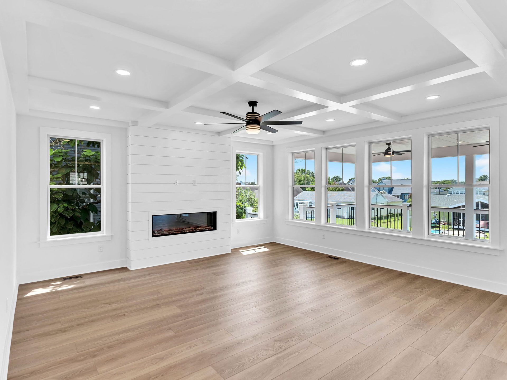 Bright, empty room with wood floors, white brick fireplace, large windows, and ceiling beams.